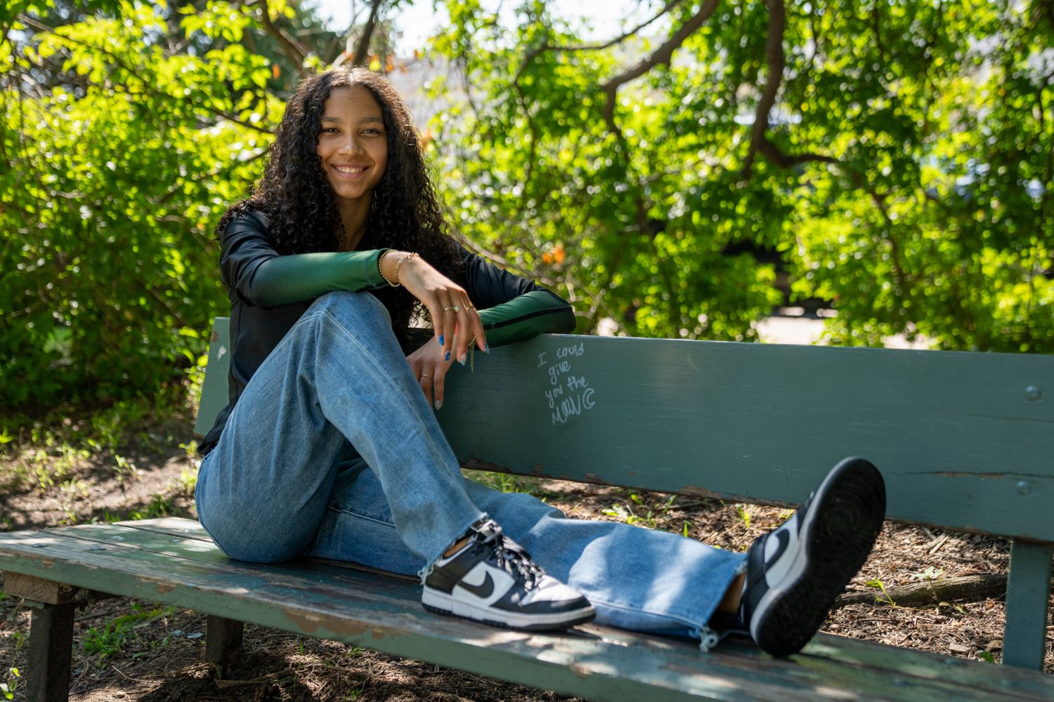 Young woman with curly hair in jeans sitting relaxed on a green park bench outdoors.
