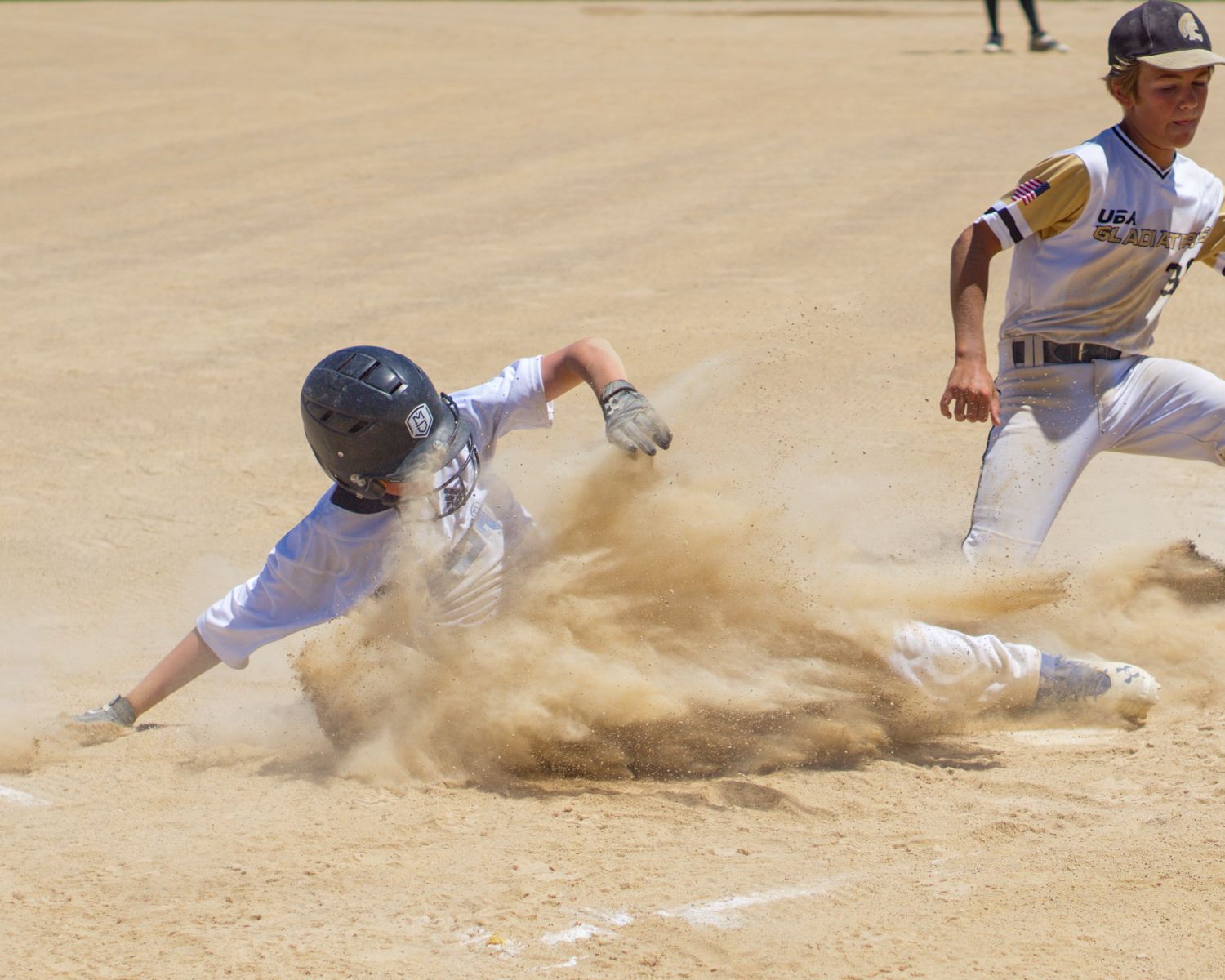 Baseball player slides into base in cloud of dust as fielder rushes to make play.