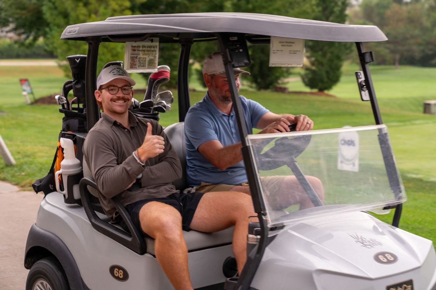 Two men smile while riding in a golf cart on a sunny golf course, one giving a thumbs up.