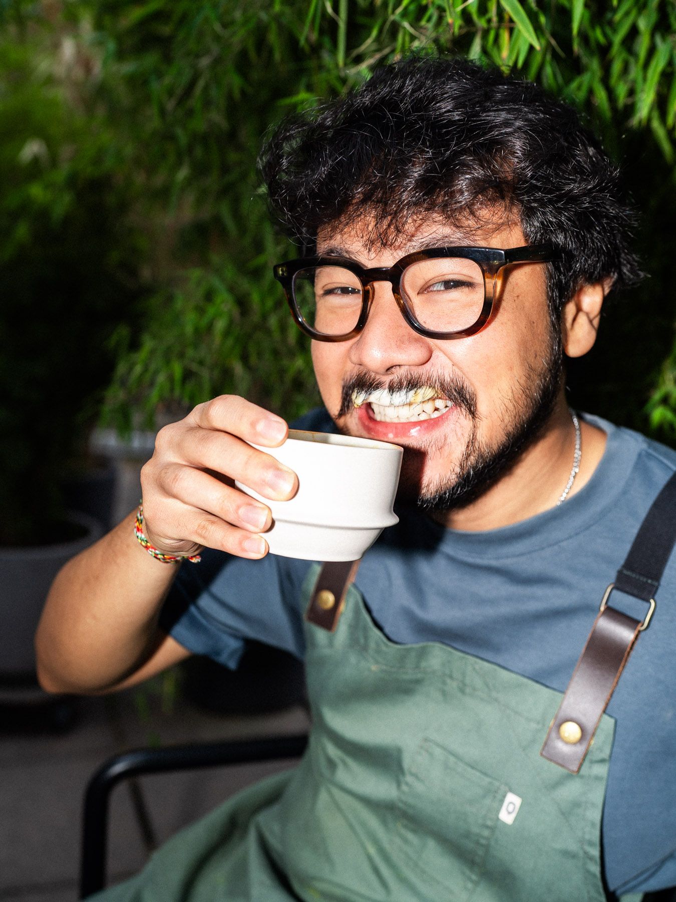 Portrait von Nico, Student und Barista aus Indonesien, mit einer Baristaschürze, während er eine Tasse Cappuccino trinkt, mit Milchbart