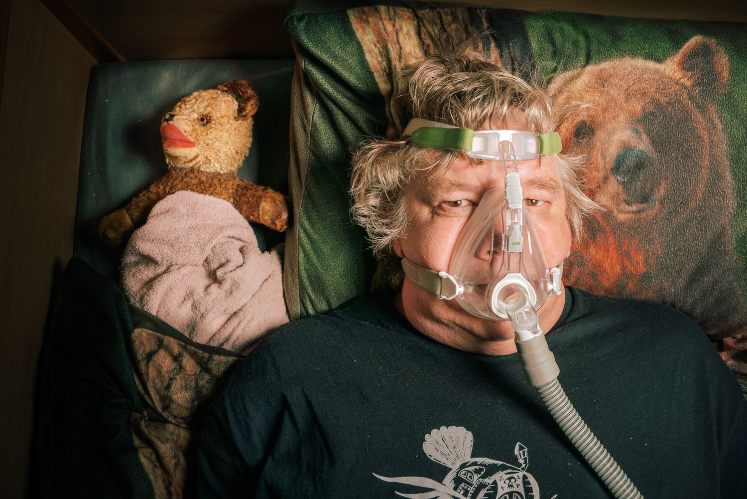 Torsten Herrmann liegt in seinem Bett mit Bärenbettwäsche, er trägt eine Schlafapoemaske. Neben ihm liegt sein Stoffbär 