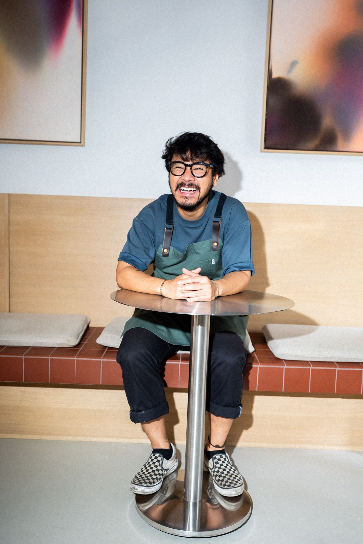 Portrait von Nico Estrada in Arbeitskleidung, lachend an einem Tisch sitzend an seinem Arbeitsplatz, dem Yuzu Cafe in Hannover