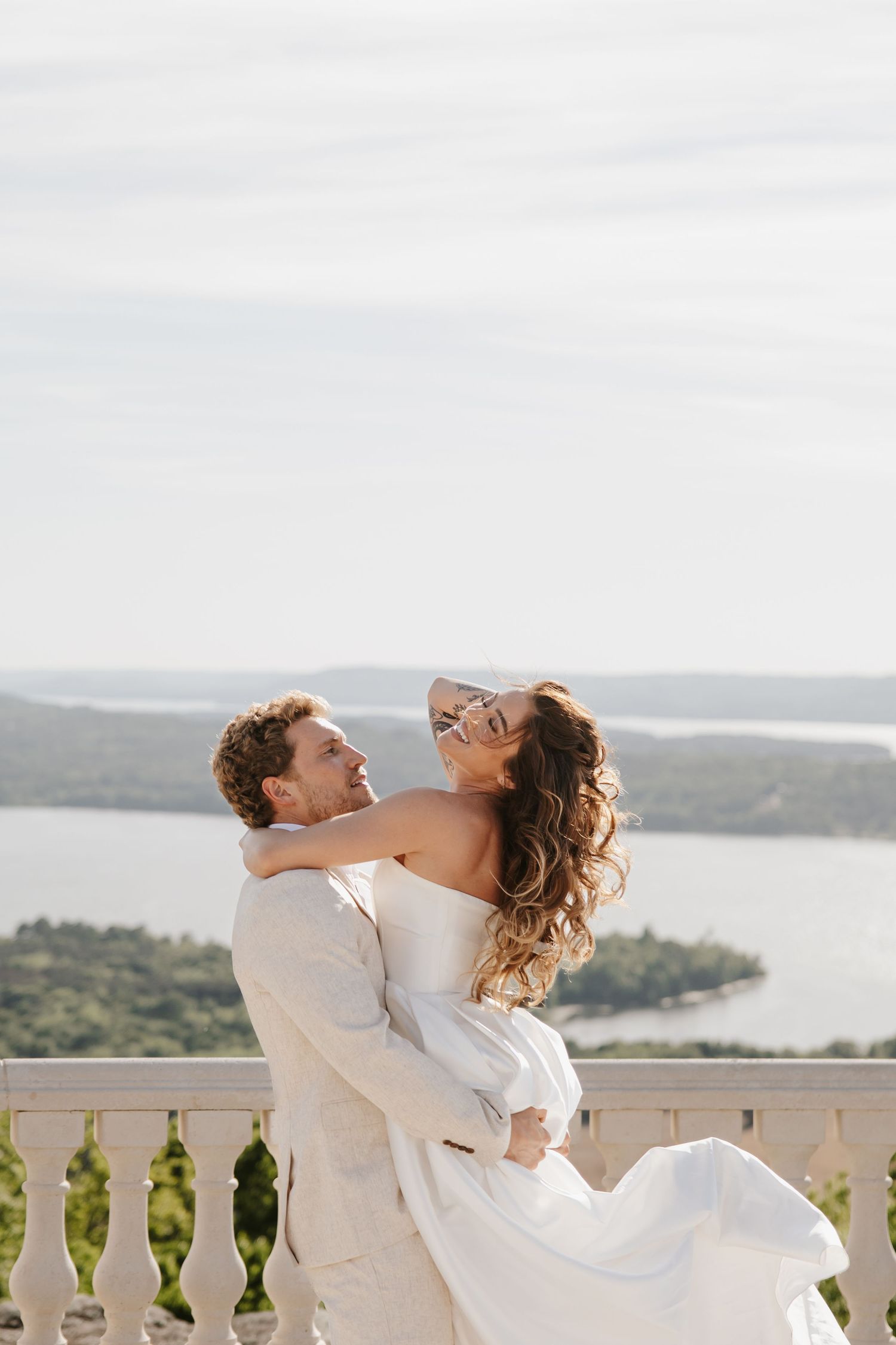 Groom lifts laughing bride in strapless gown on balcony overlooking scenic lake and green hills.