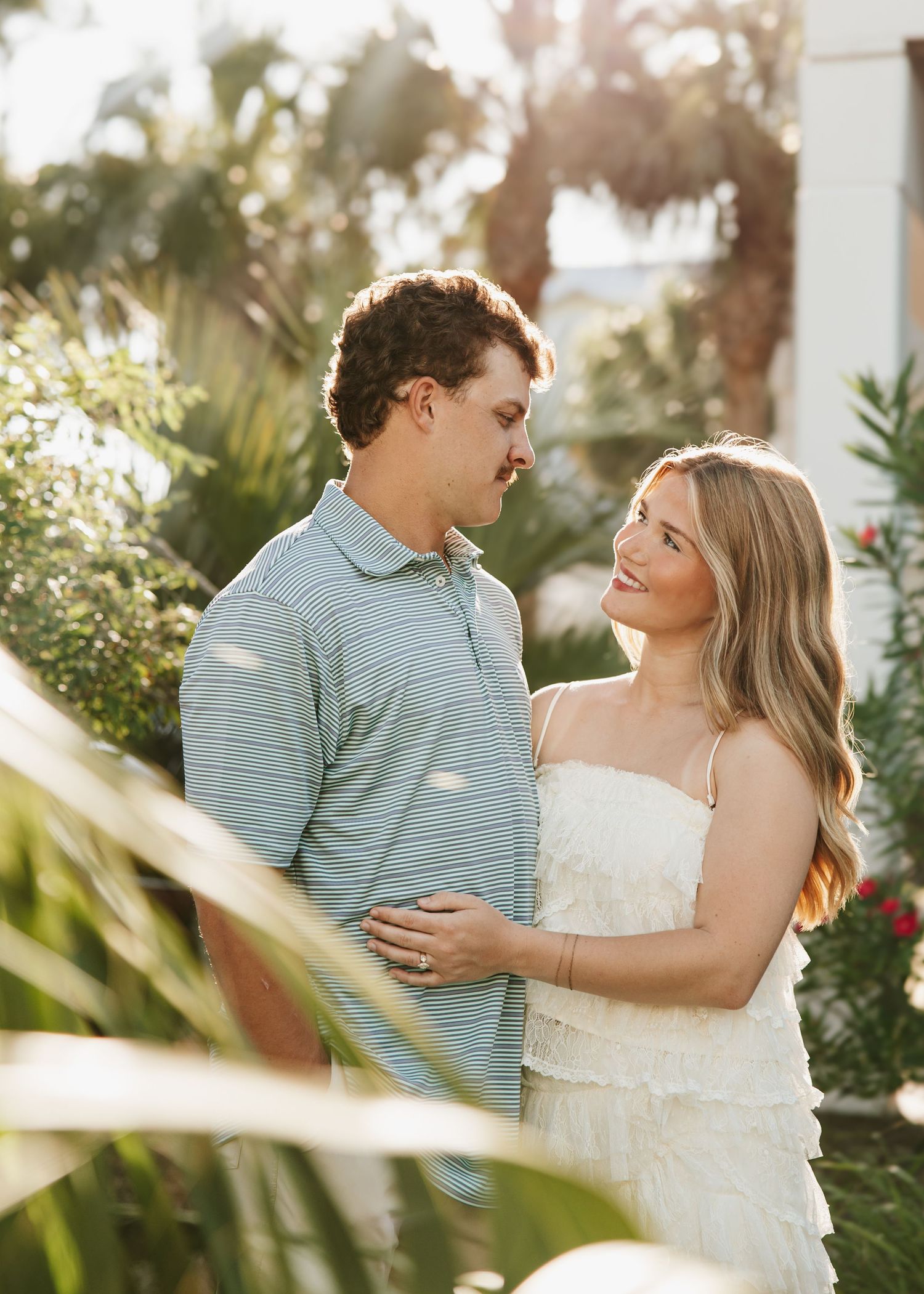 Smiling couple shares a tender moment surrounded by lush tropical greenery in warm golden sunlight.