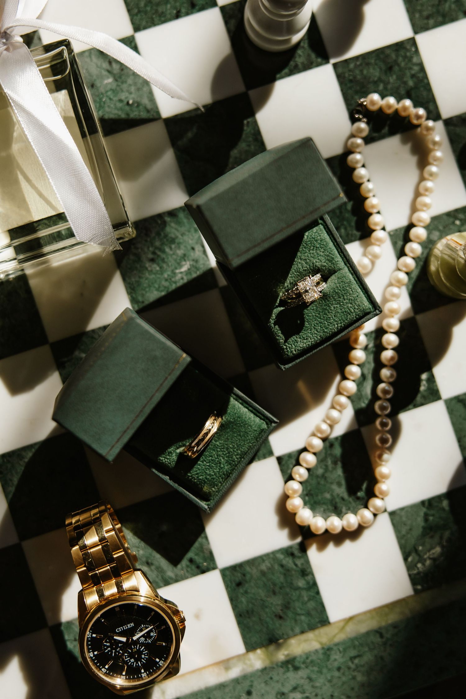 Wedding flat lay with green velvet ring boxes, pearl necklace, gold watch, and perfume on marble.