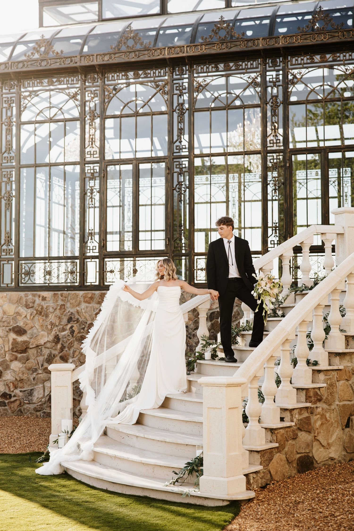 Bride and groom descend ornate white stairs before a stunning Victorian-style glass greenhouse structure.