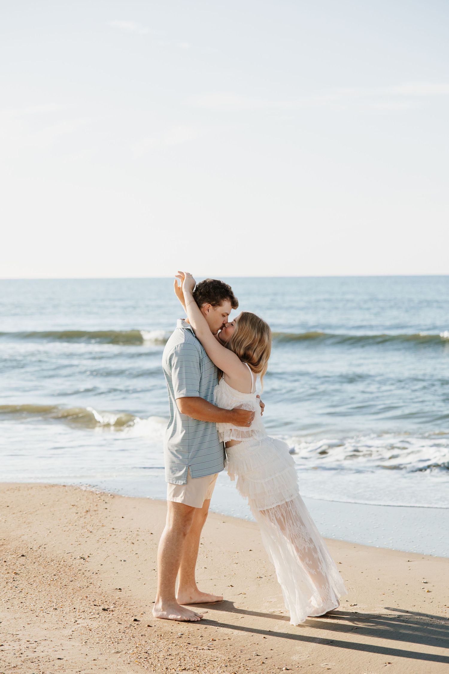 Couple embraces and kisses barefoot on a sunny sandy beach with ocean waves behind them.
