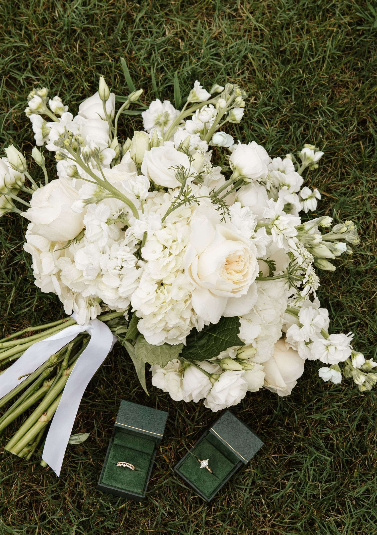 White wedding bouquet of peonies and roses rests on grass beside two green velvet ring boxes.