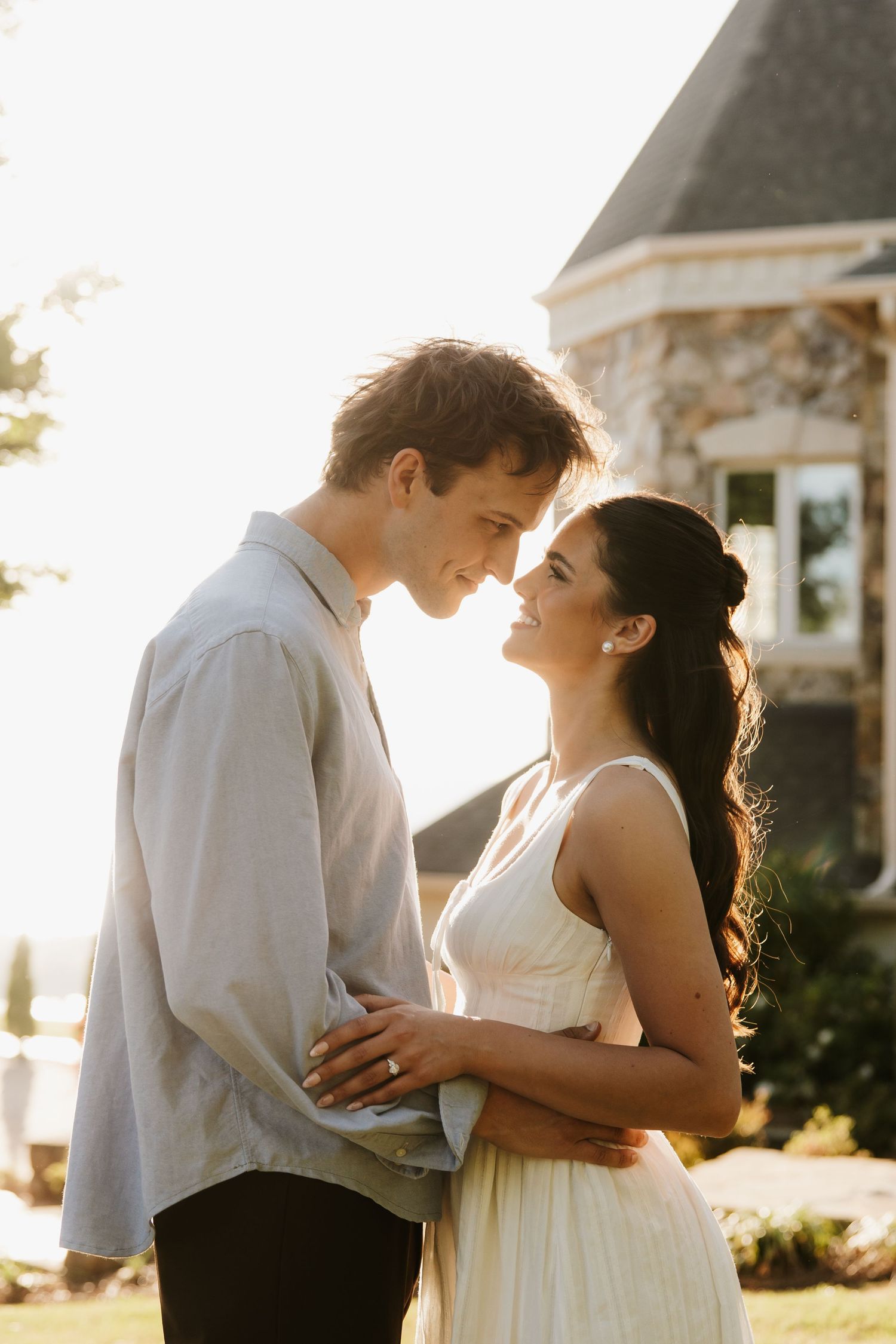 Couple in golden sunset light embrace nose-to-nose outside a stone building in casual wedding attire.