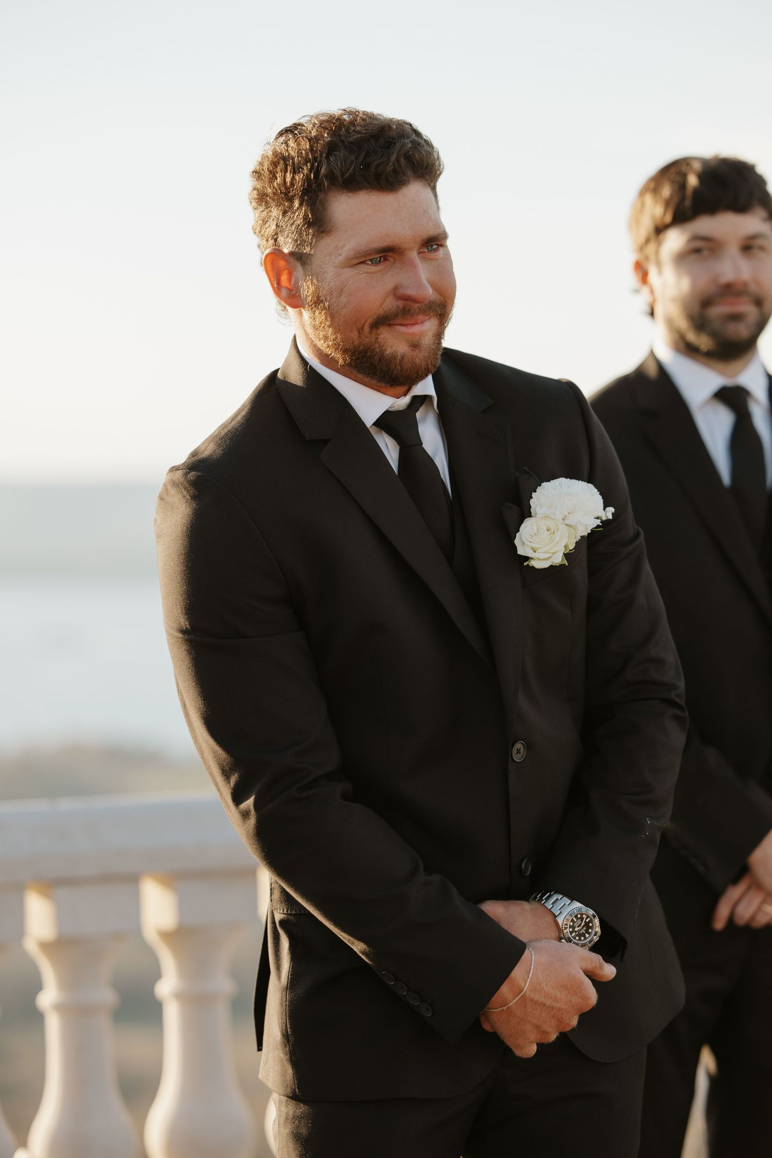 Groom smiling in black suit with white boutonniere at outdoor wedding ceremony.