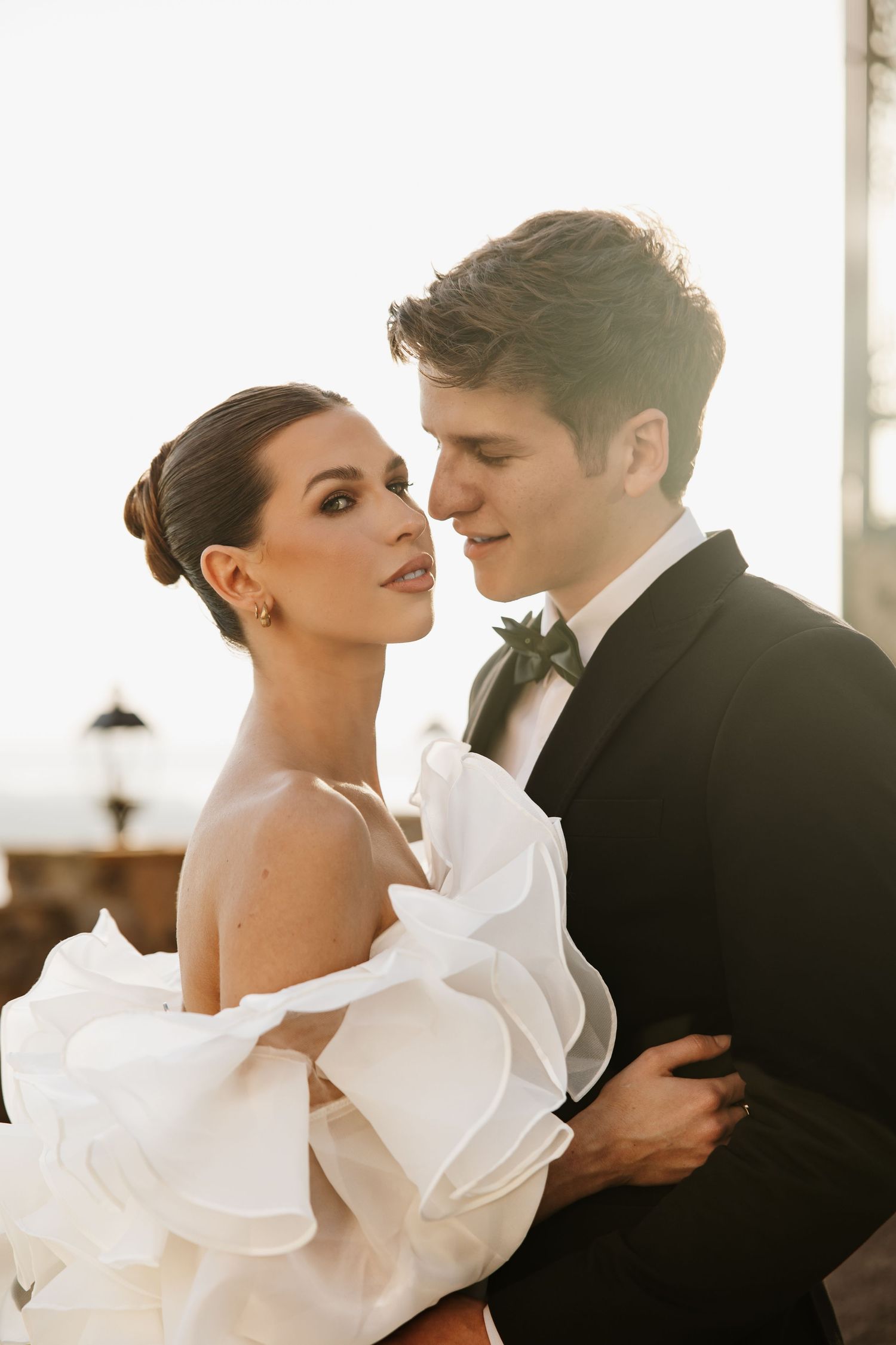 Bride in off-shoulder white ruffled gown and groom in black tuxedo embrace romantically at golden hour.