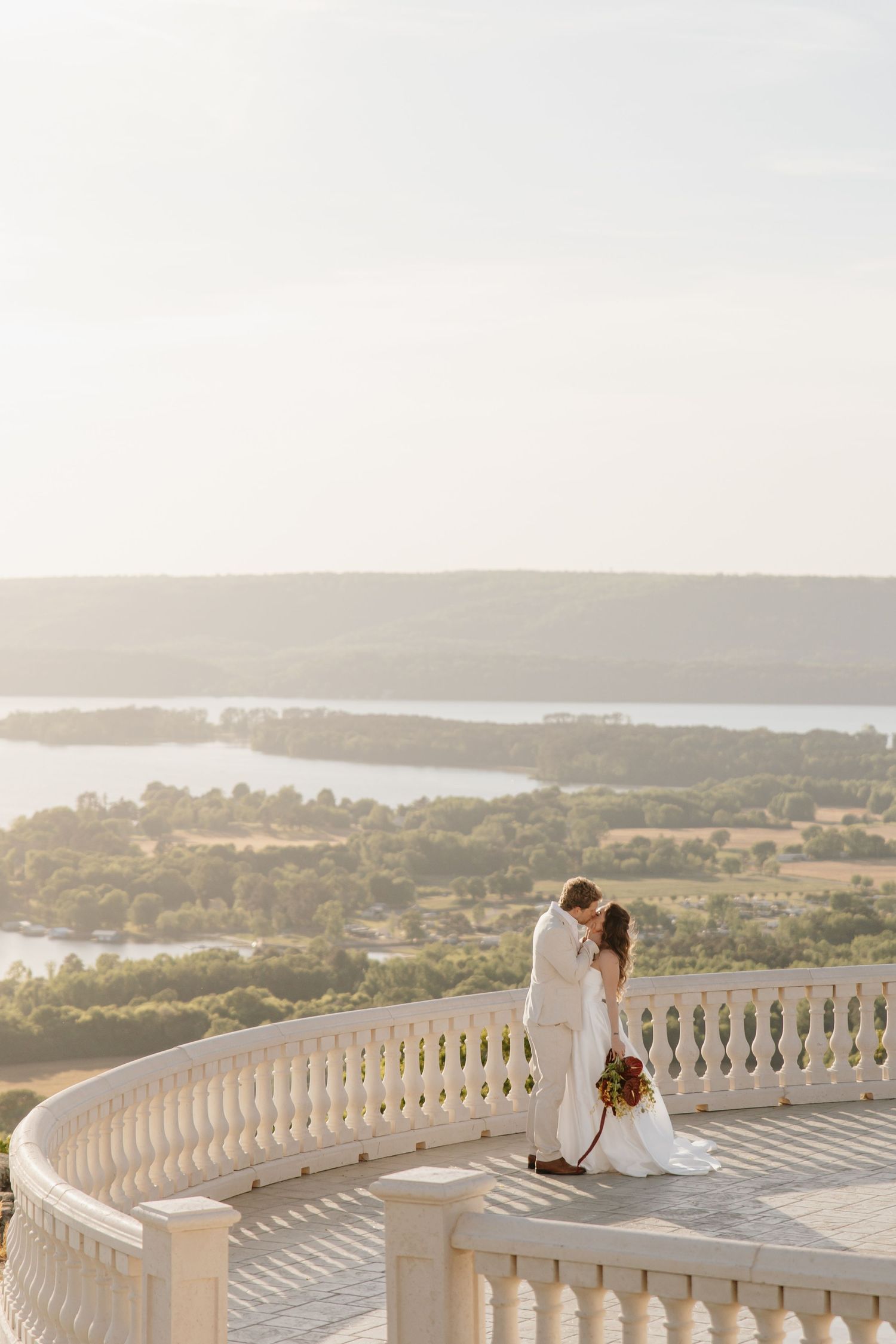 Romantic couple shares a kiss on a curved balcony overlooking a breathtaking river landscape at sunset.