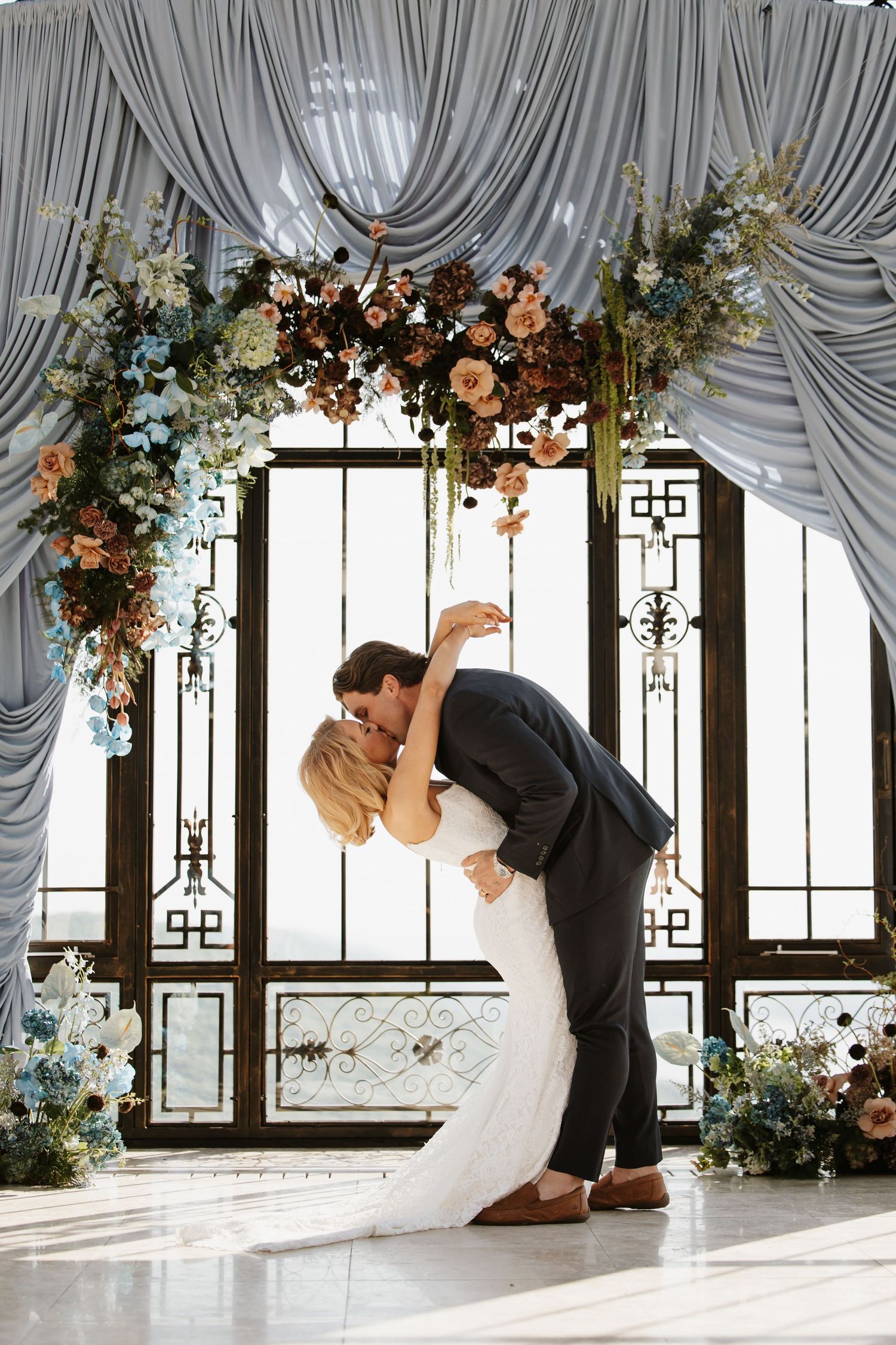 Bride and groom share a dip kiss under a floral arch with blue draping and colorful blooms.