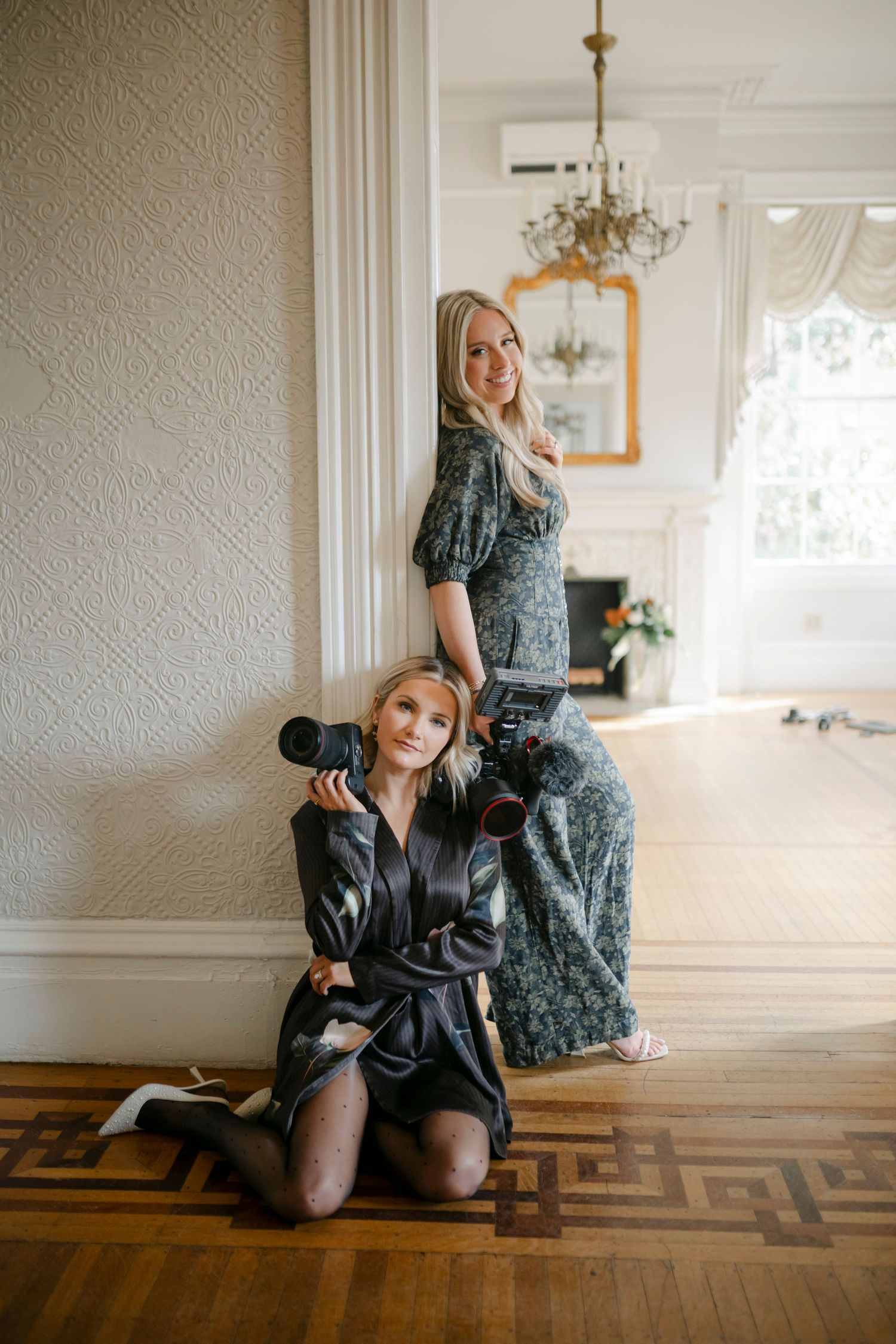 Two female wedding photographers pose with cameras in elegant white room with chandelier and hardwood floors.