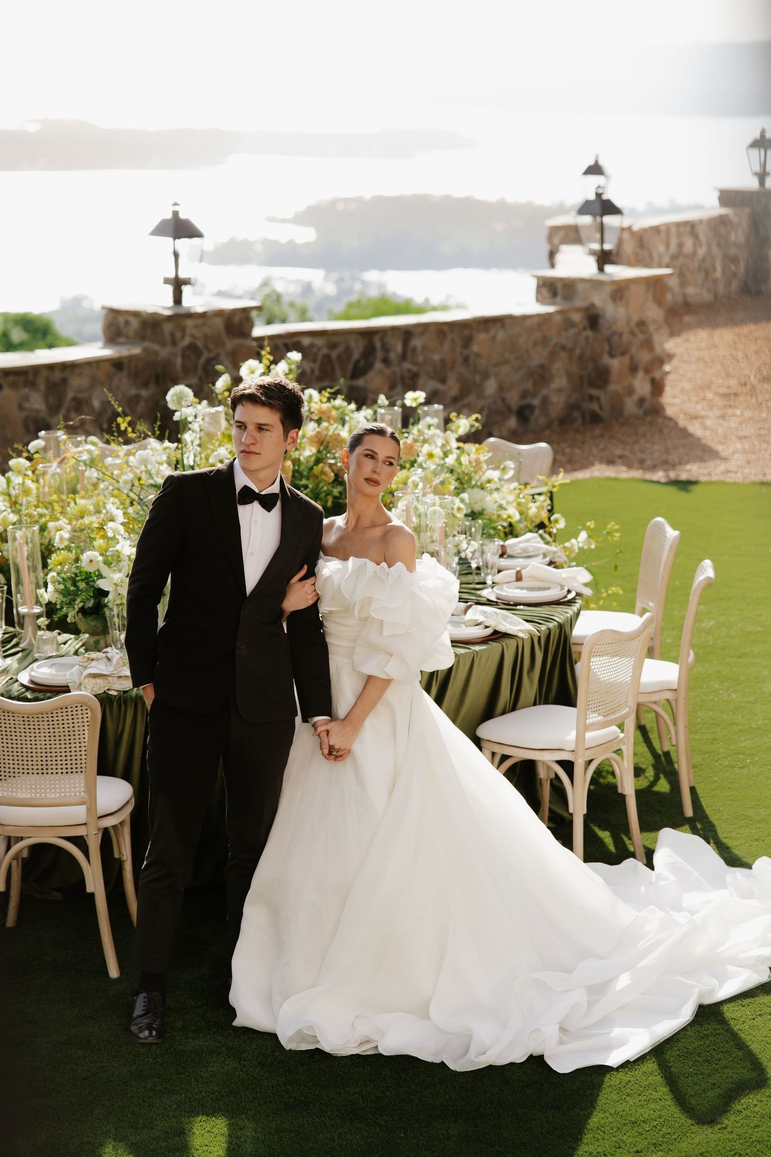 Elegant bride in off-shoulder white gown and groom in black tuxedo pose beside floral wedding table with scenic water view.