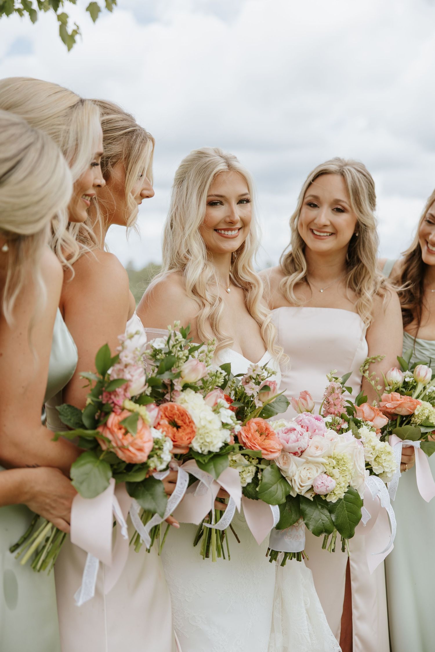 Smiling bride in white gown surrounded by bridesmaids in sage green dresses holding colorful bouquets.