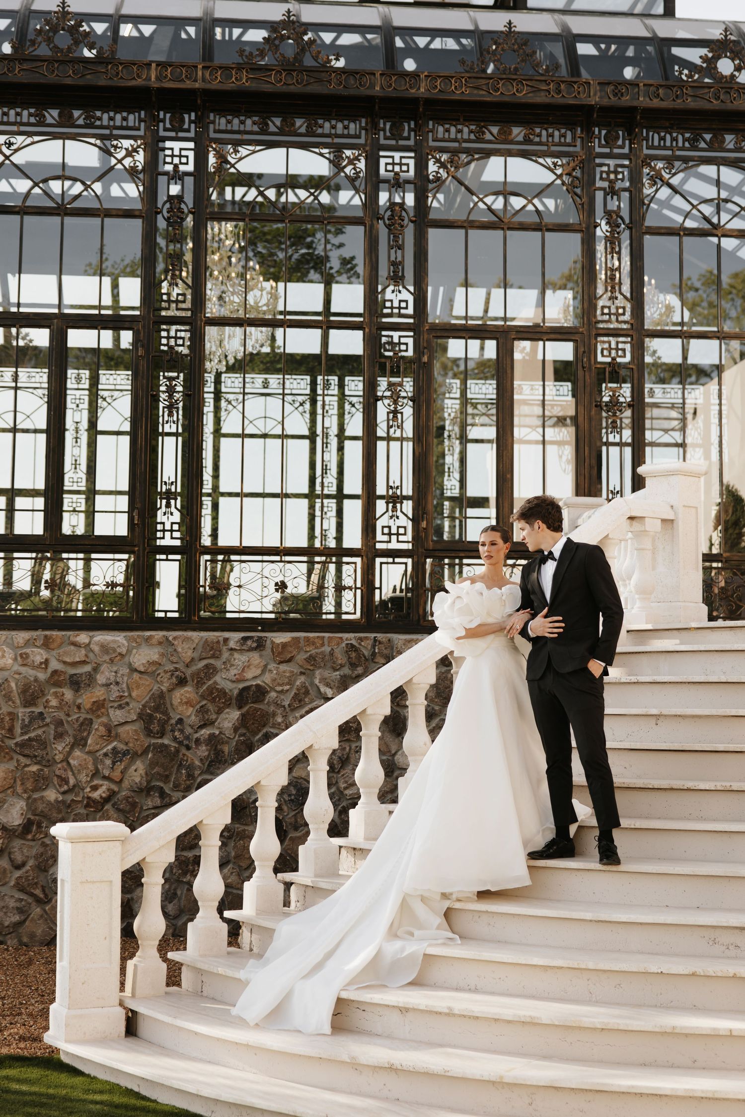 Bride and groom in elegant attire pose on white marble staircase before ornate glass greenhouse.