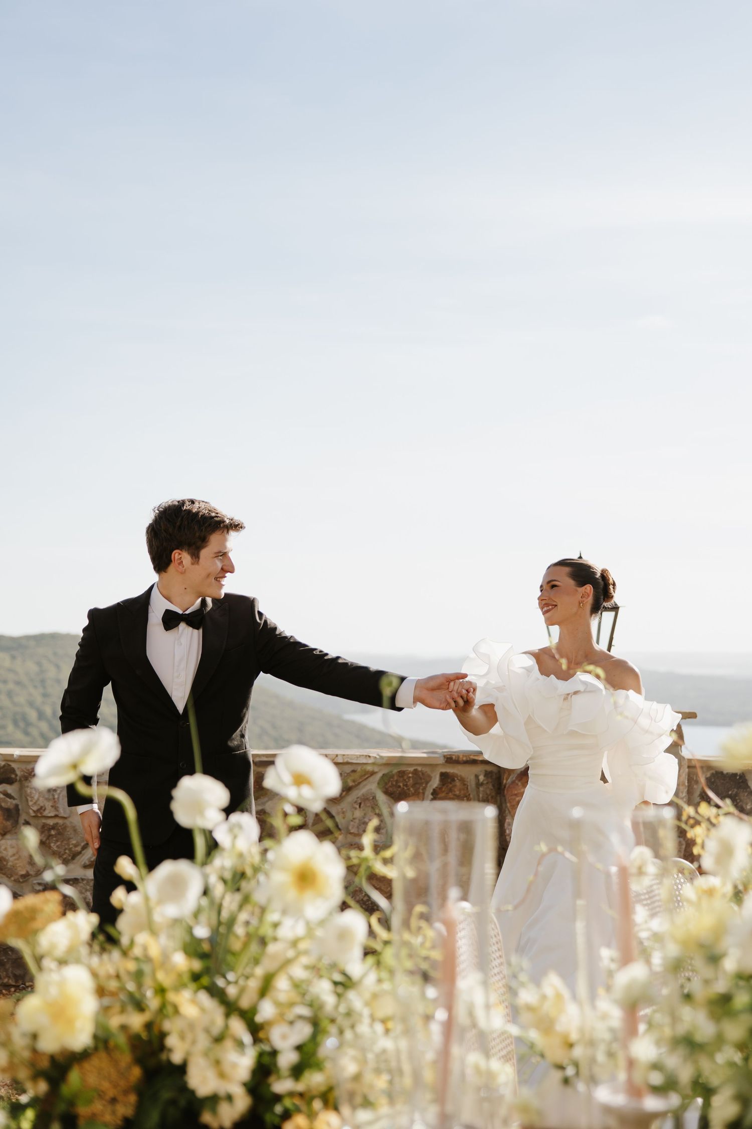 Joyful bride and groom hold hands laughing at outdoor reception table with scenic hilltop view.