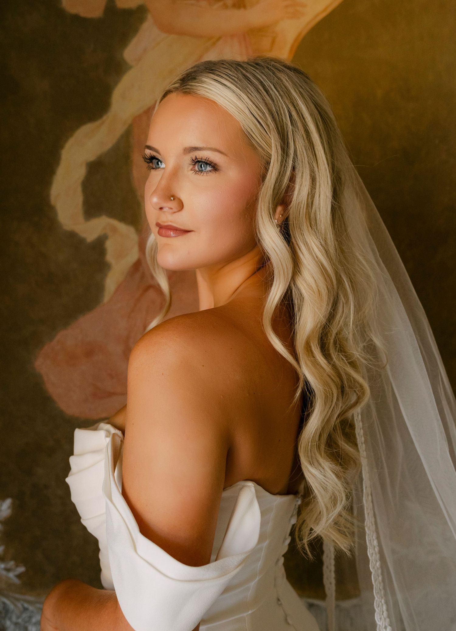 Blonde bride in an off-shoulder white gown with flowing veil poses before a painted wall.
