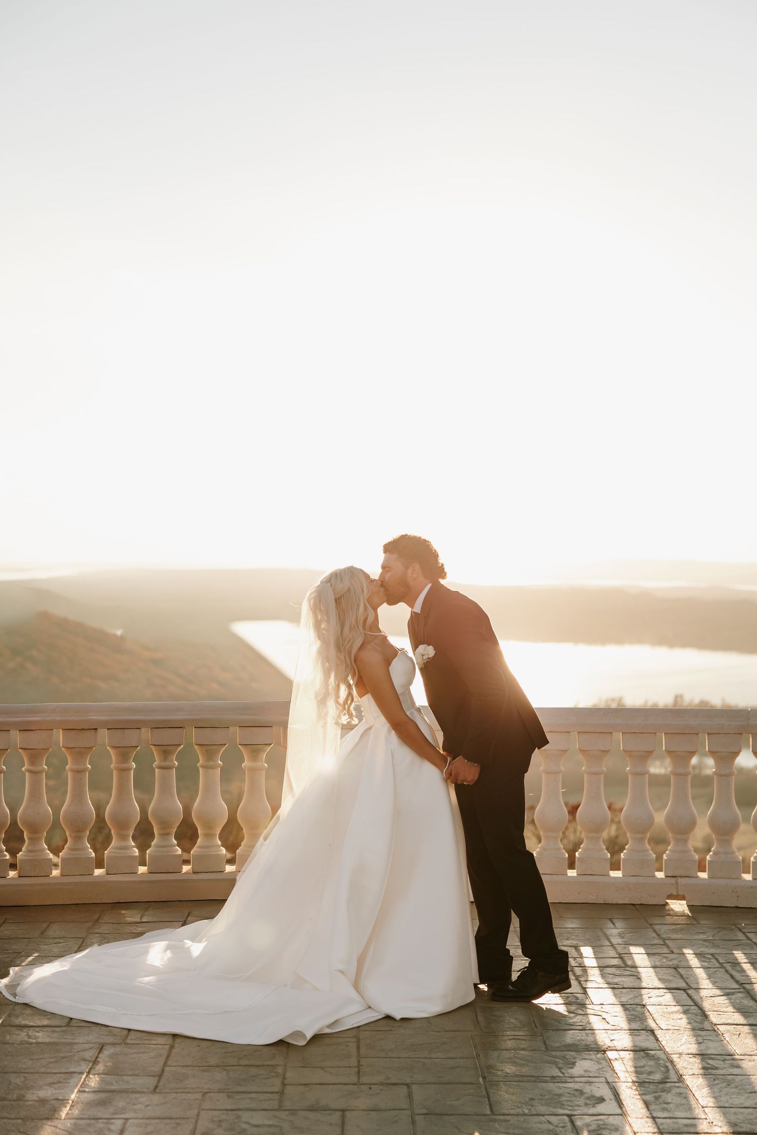 Bride and groom share a kiss on a balcony at golden hour with a scenic view.