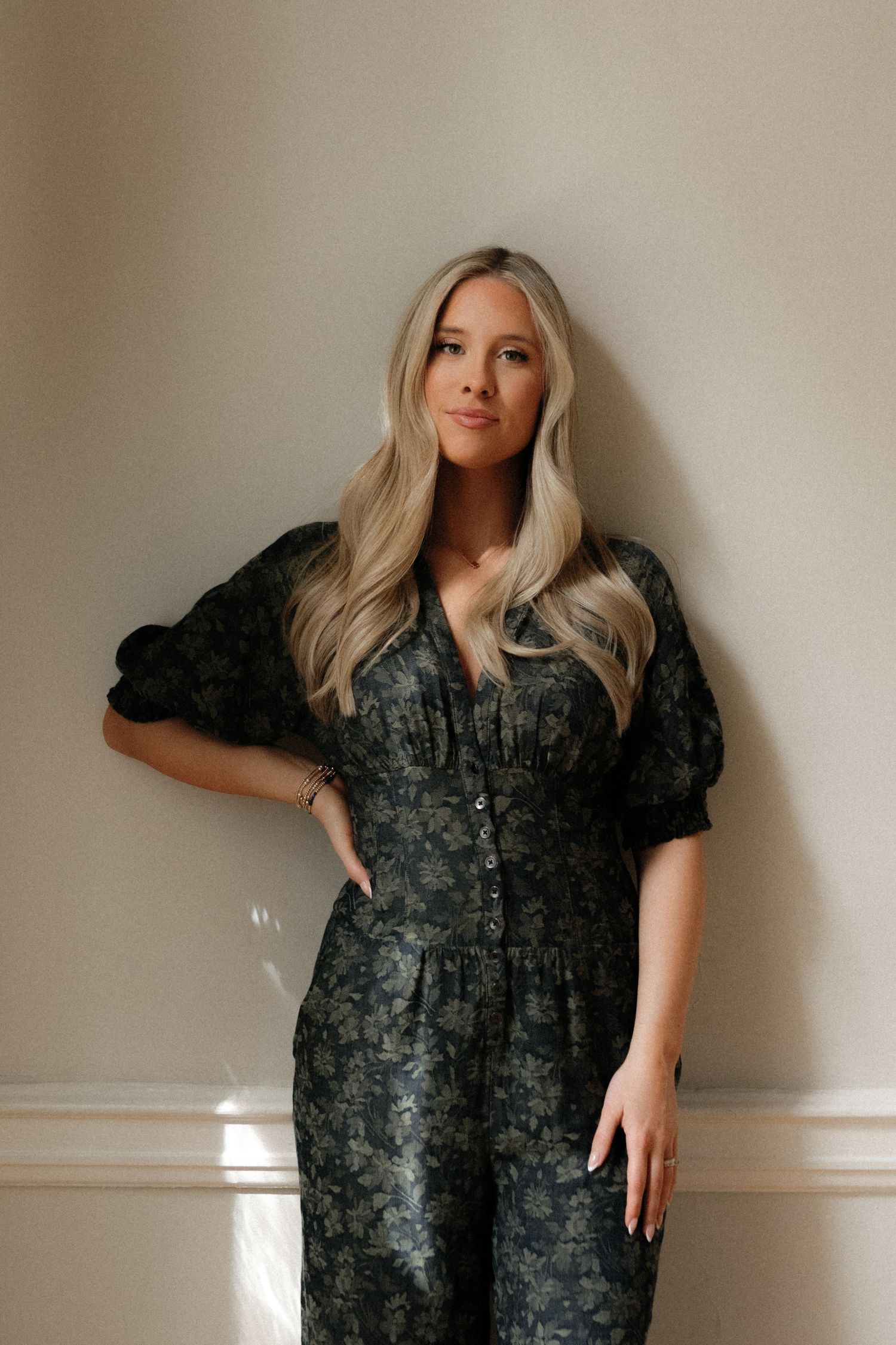 Blonde woman in a dark floral button-front midi dress poses confidently against a light wall.