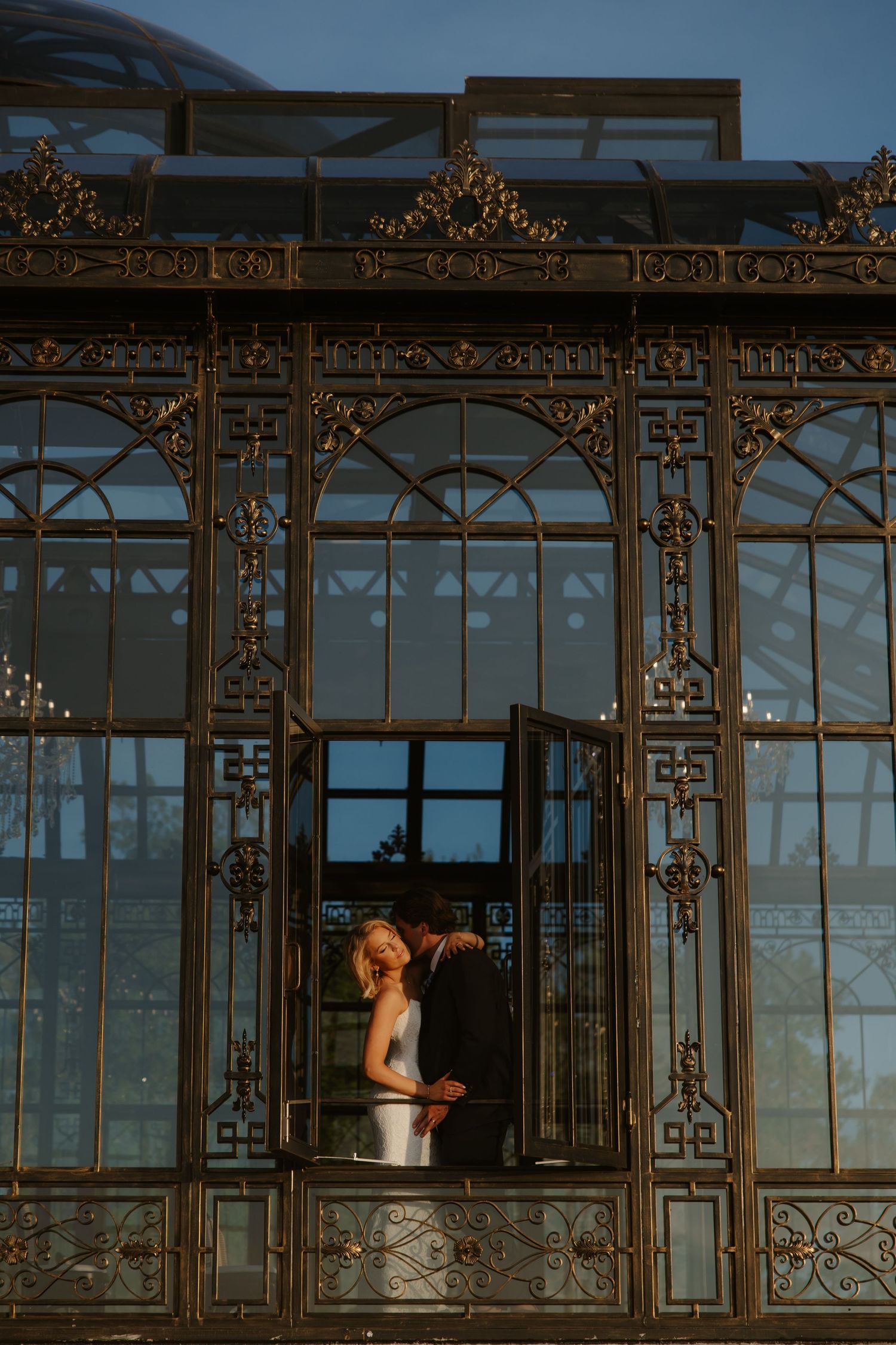 Bride and groom embrace romantically inside an ornate golden glass conservatory at dusk.