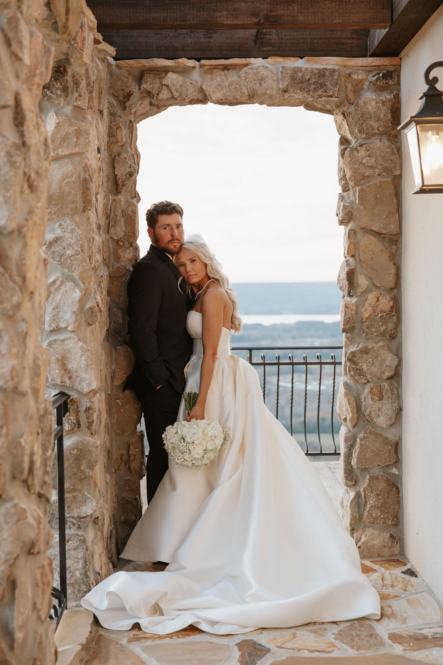 Bride and groom posing in stone archway with scenic water view behind them.