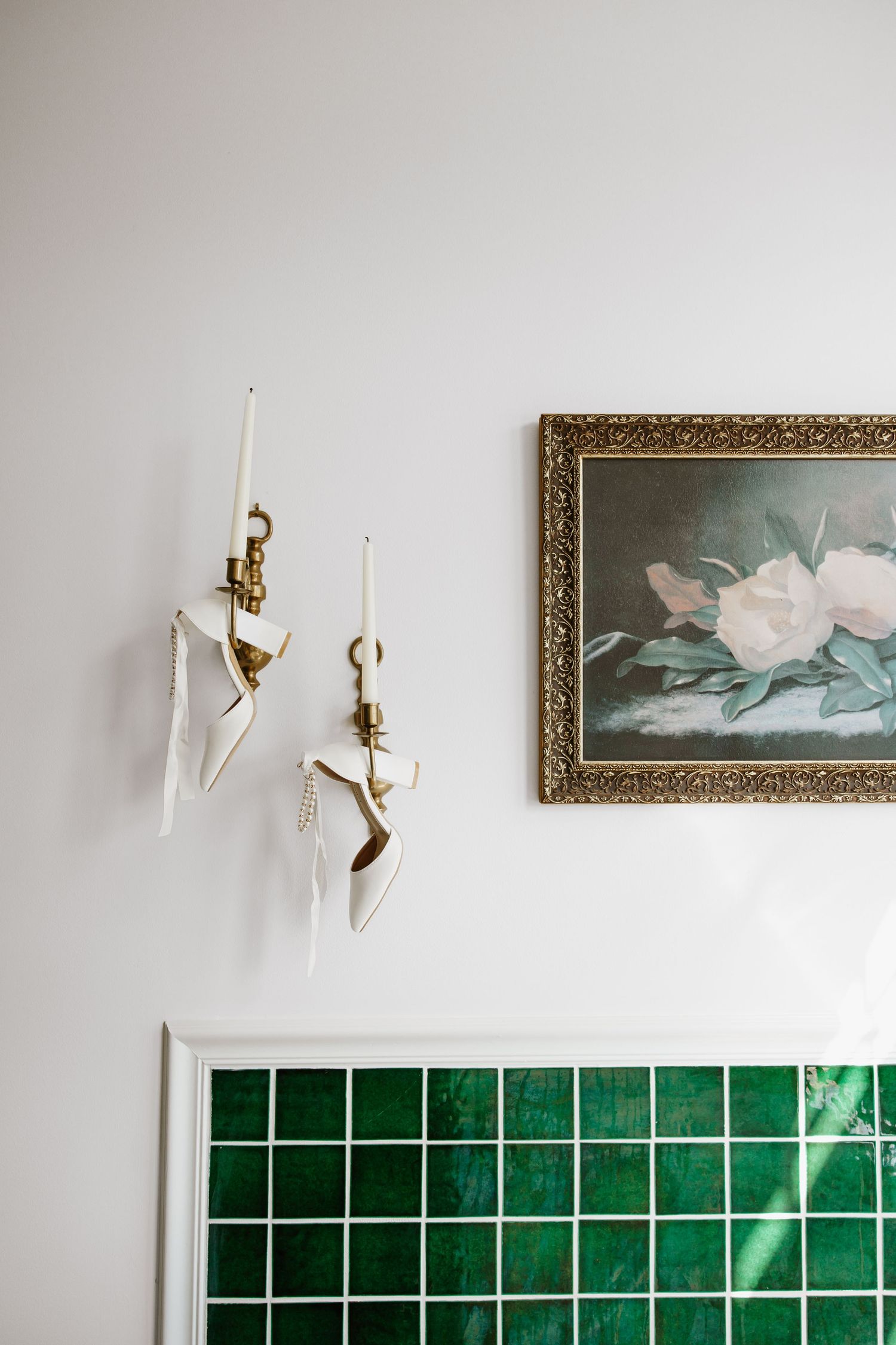 White bridal heels hang from brass wall sconces beside a floral painting above green tiled wall.