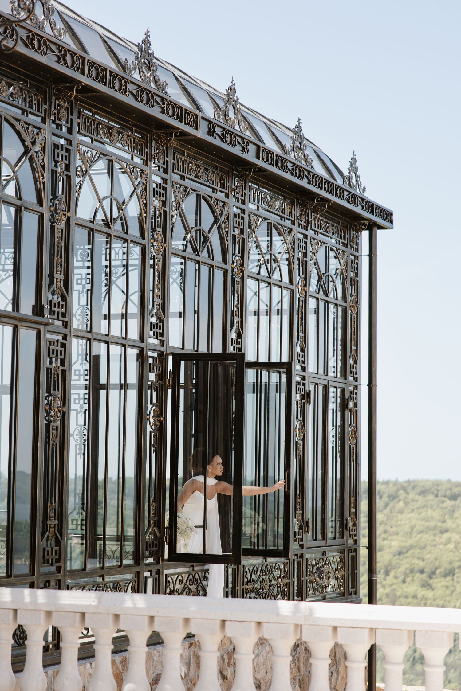 Bride in white gown standing inside an ornate black iron Victorian greenhouse structure outdoors.