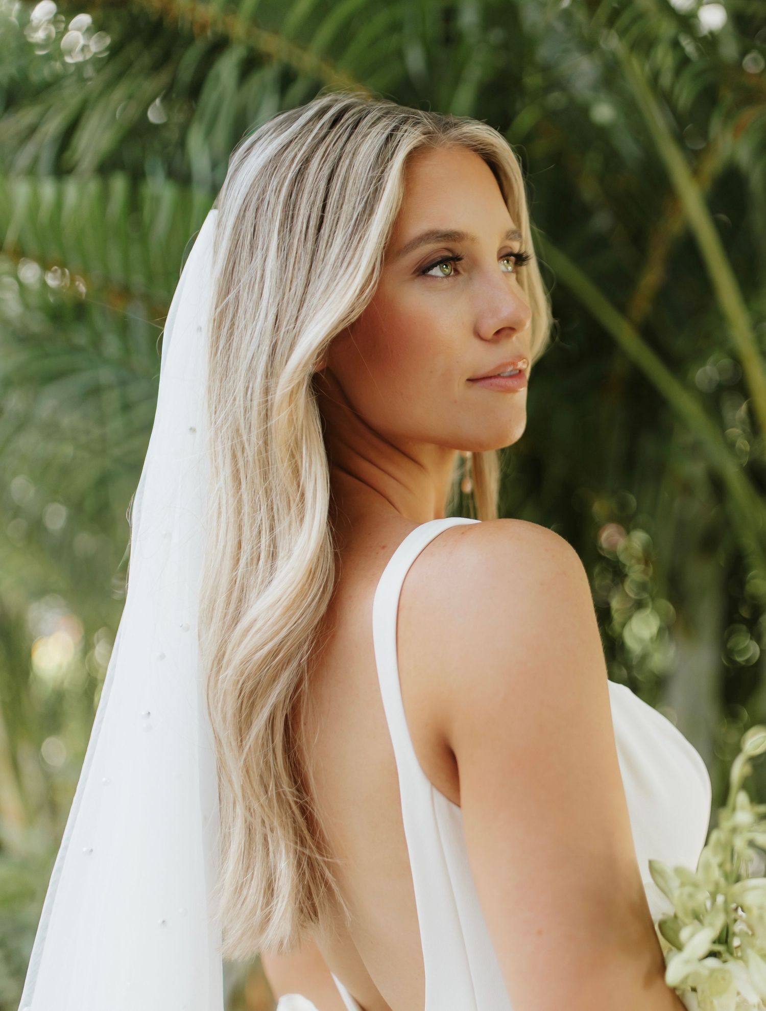 Blonde bride in a white gown and veil stands among tropical palm trees outdoors.