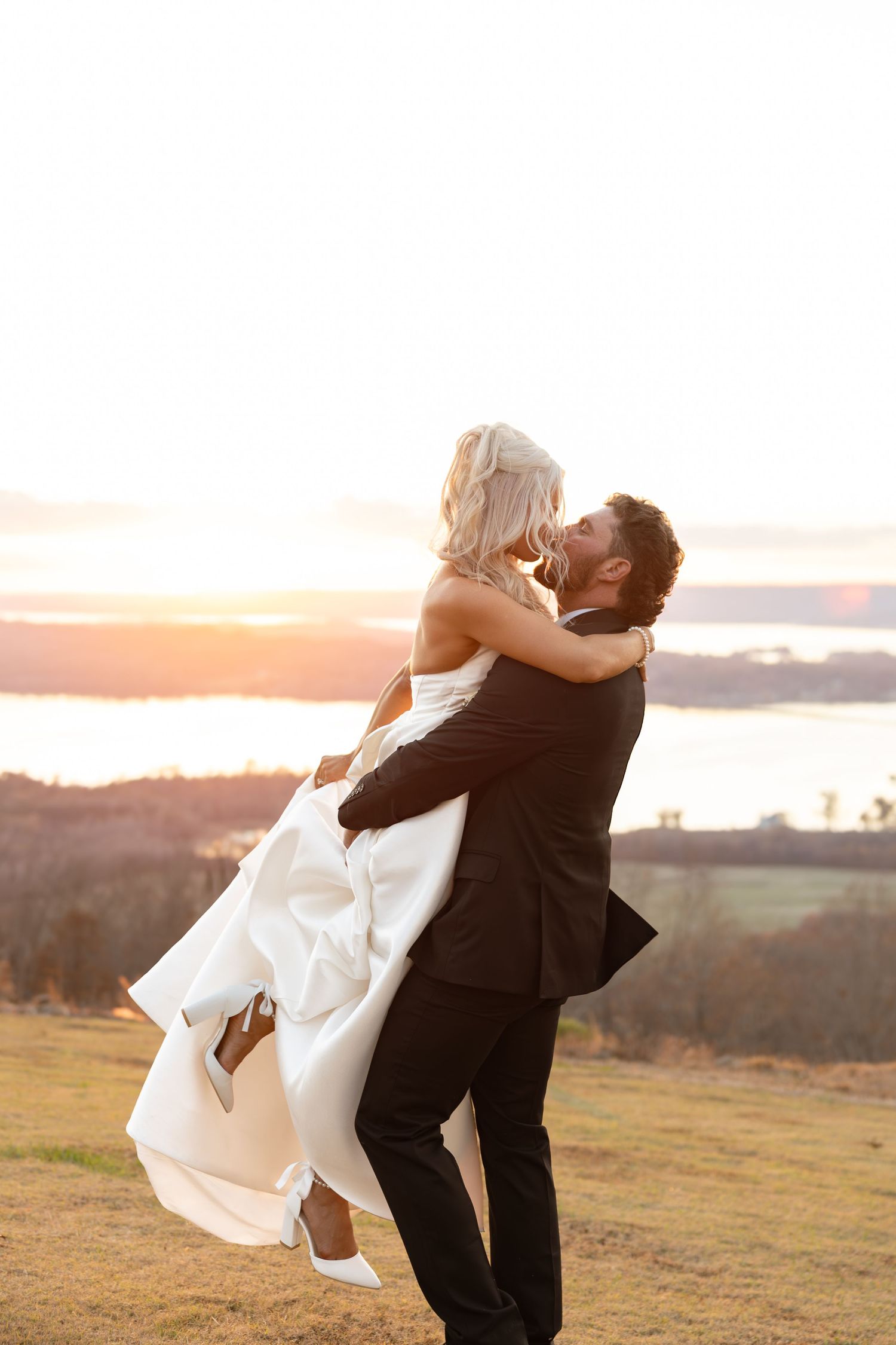 Groom lifting bride at sunset with scenic lake and golden sky in background.