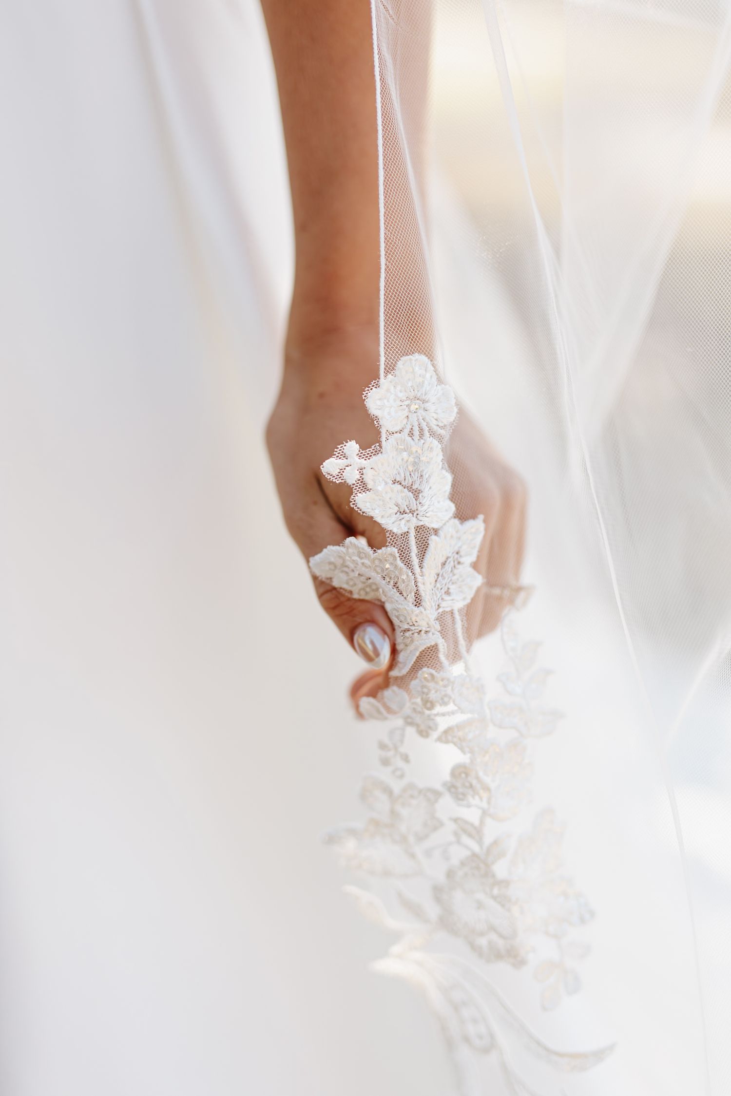 Bride holding delicate lace-trimmed wedding veil with floral embroidery detail in soft white light.