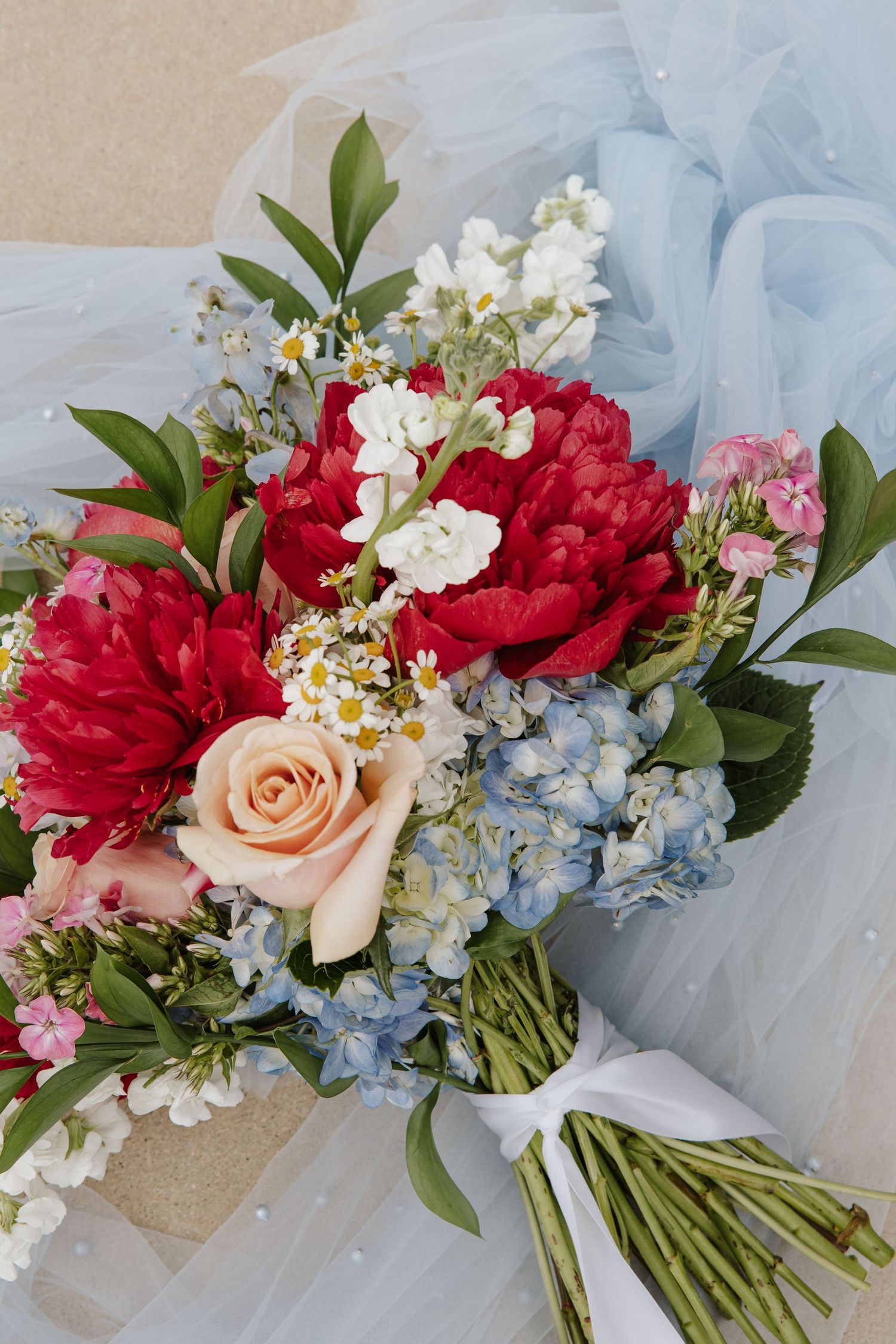 Colorful bridal bouquet with red peonies, peach roses, blue hydrangeas, and daisies on light blue tulle.