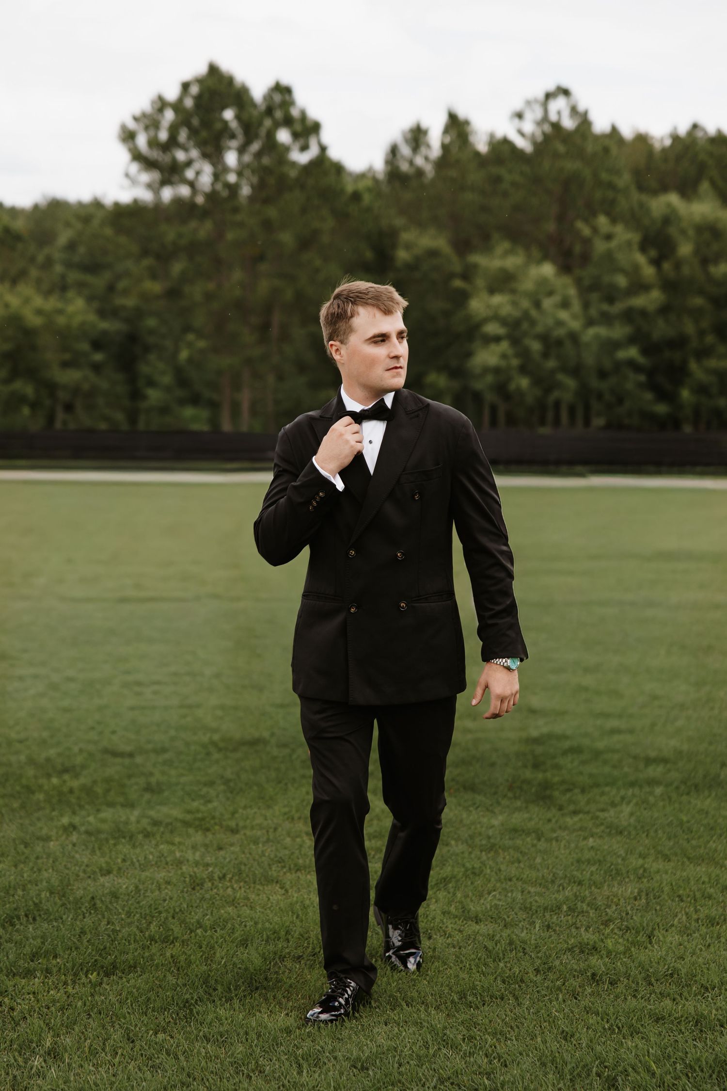 Groom in black tuxedo adjusting bow tie while walking across green lawn outdoors.