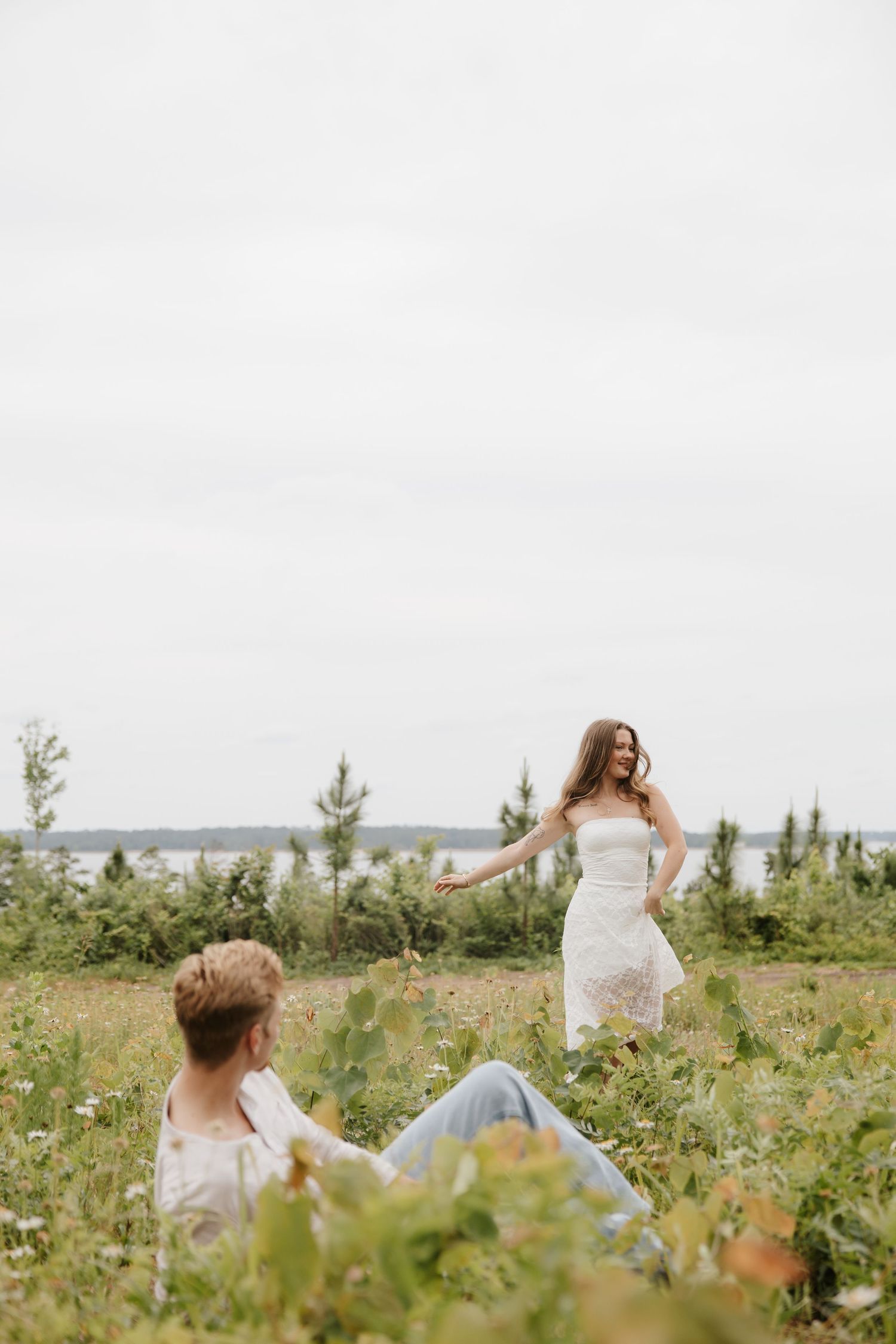 Bride in white lace dress walks toward partner sitting in a wildflower meadow under cloudy skies.
