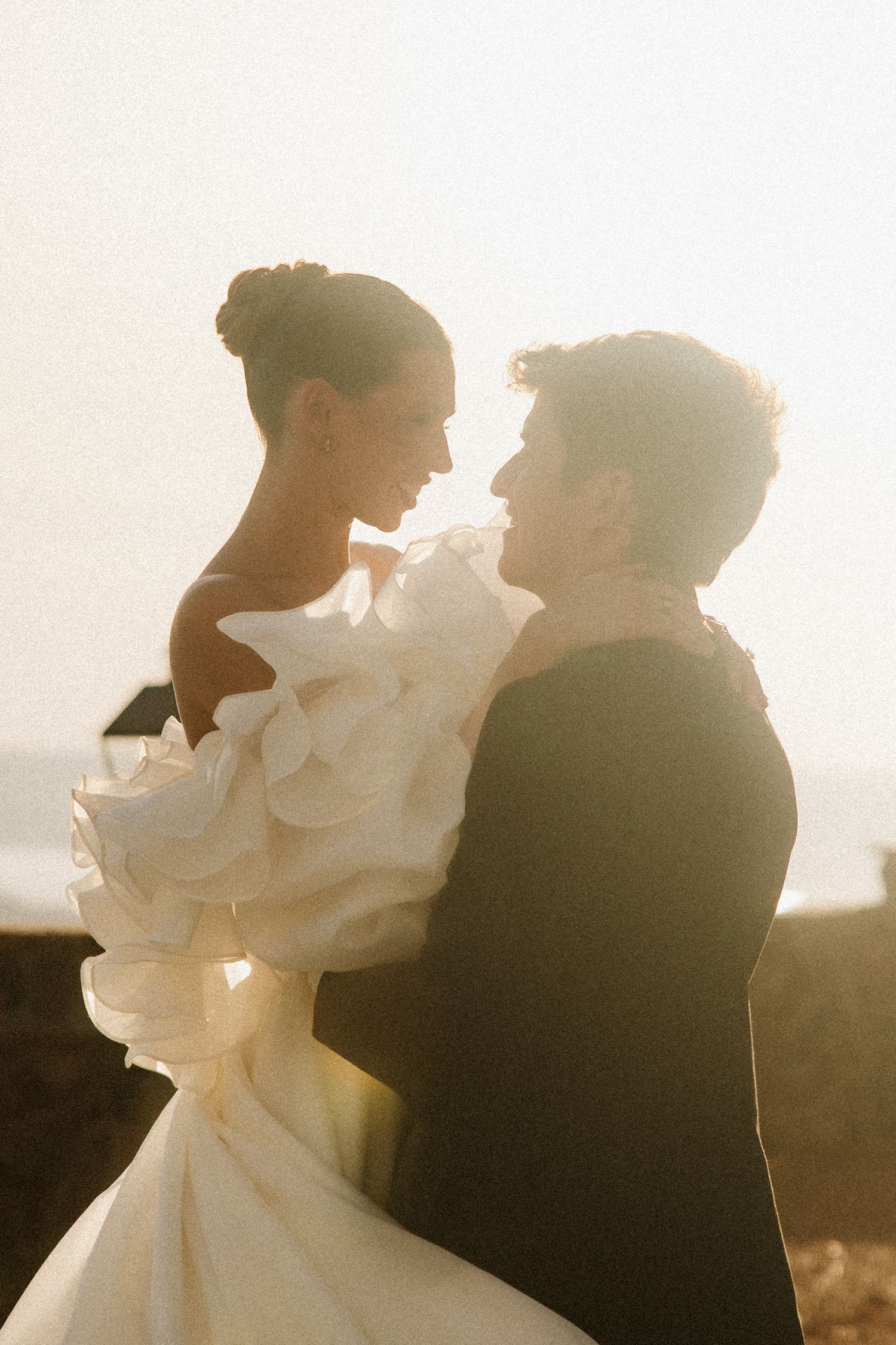 Bride and groom embrace in golden backlight, her ruffled off-shoulder gown glowing against a sunlit coastal backdrop.