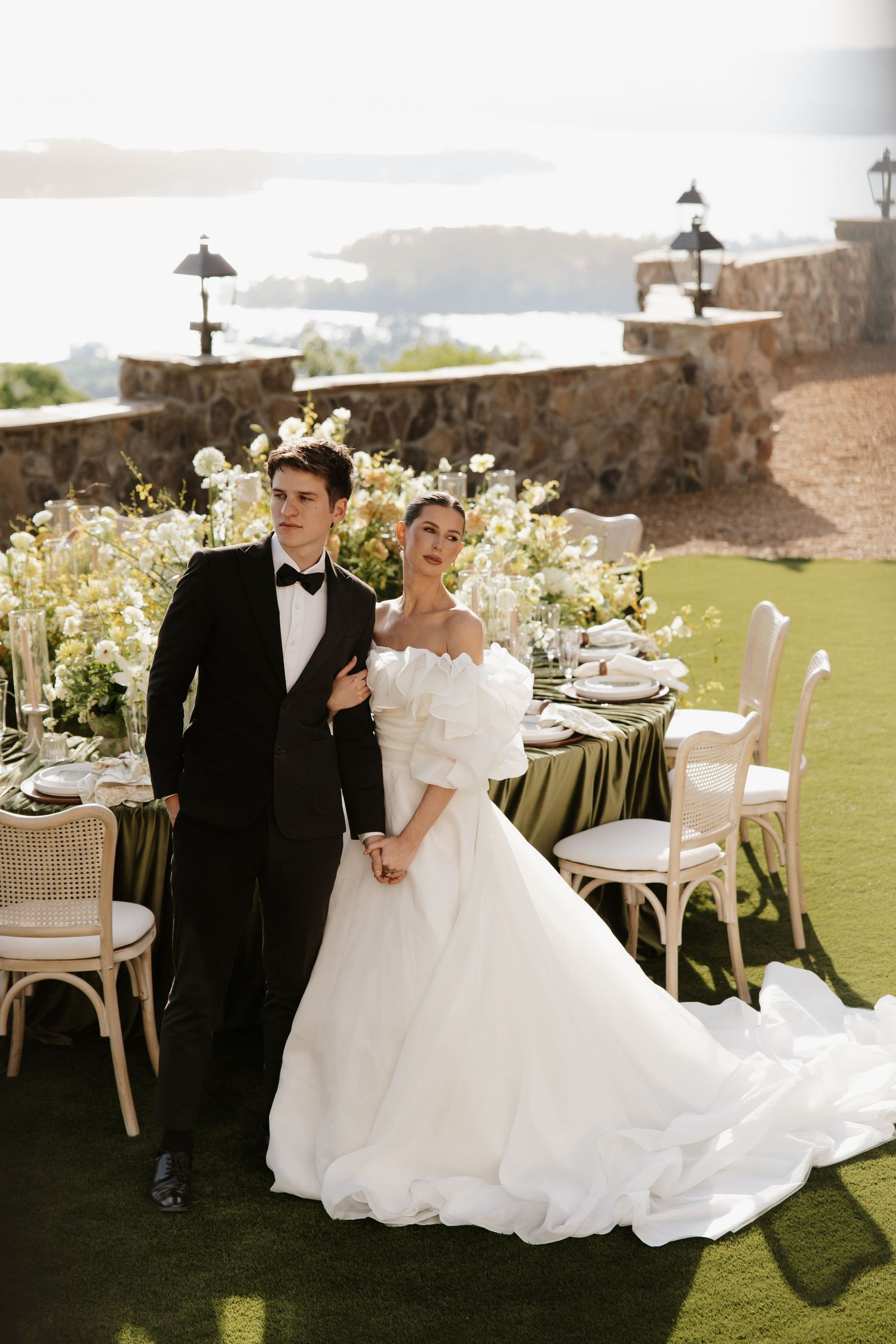 Elegant bride and groom pose beside an outdoor reception table with floral arrangements and scenic water views.