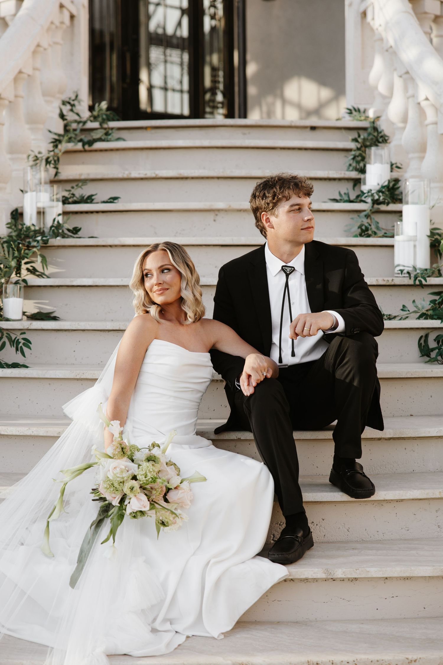Bride and groom sit on white decorated steps, bride holding pink and white bouquet in strapless ballgown.