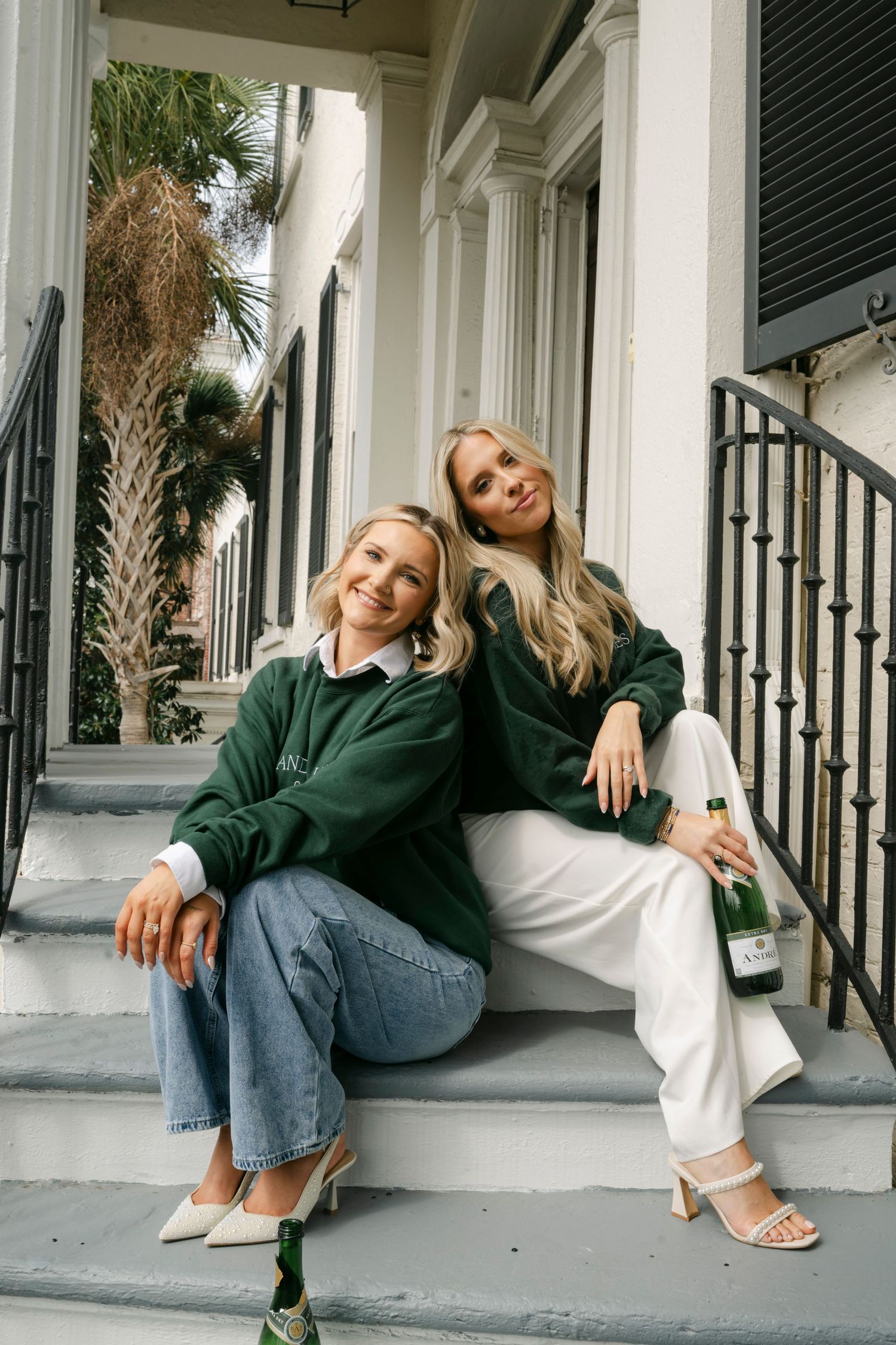 Two blonde women in matching green sweaters sit on porch steps holding a champagne bottle.