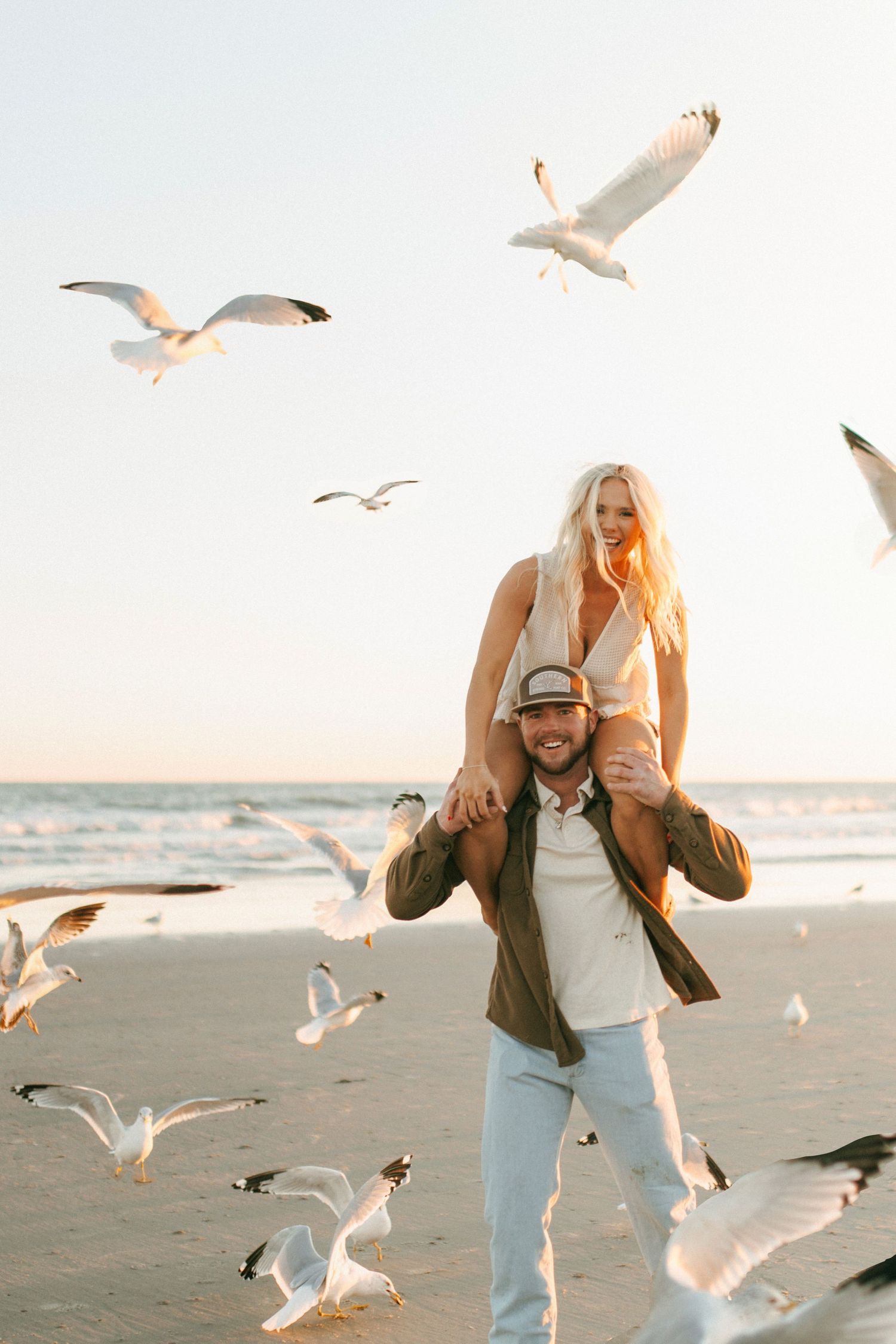 Couple laughing on beach with seagulls flying around them at golden hour.