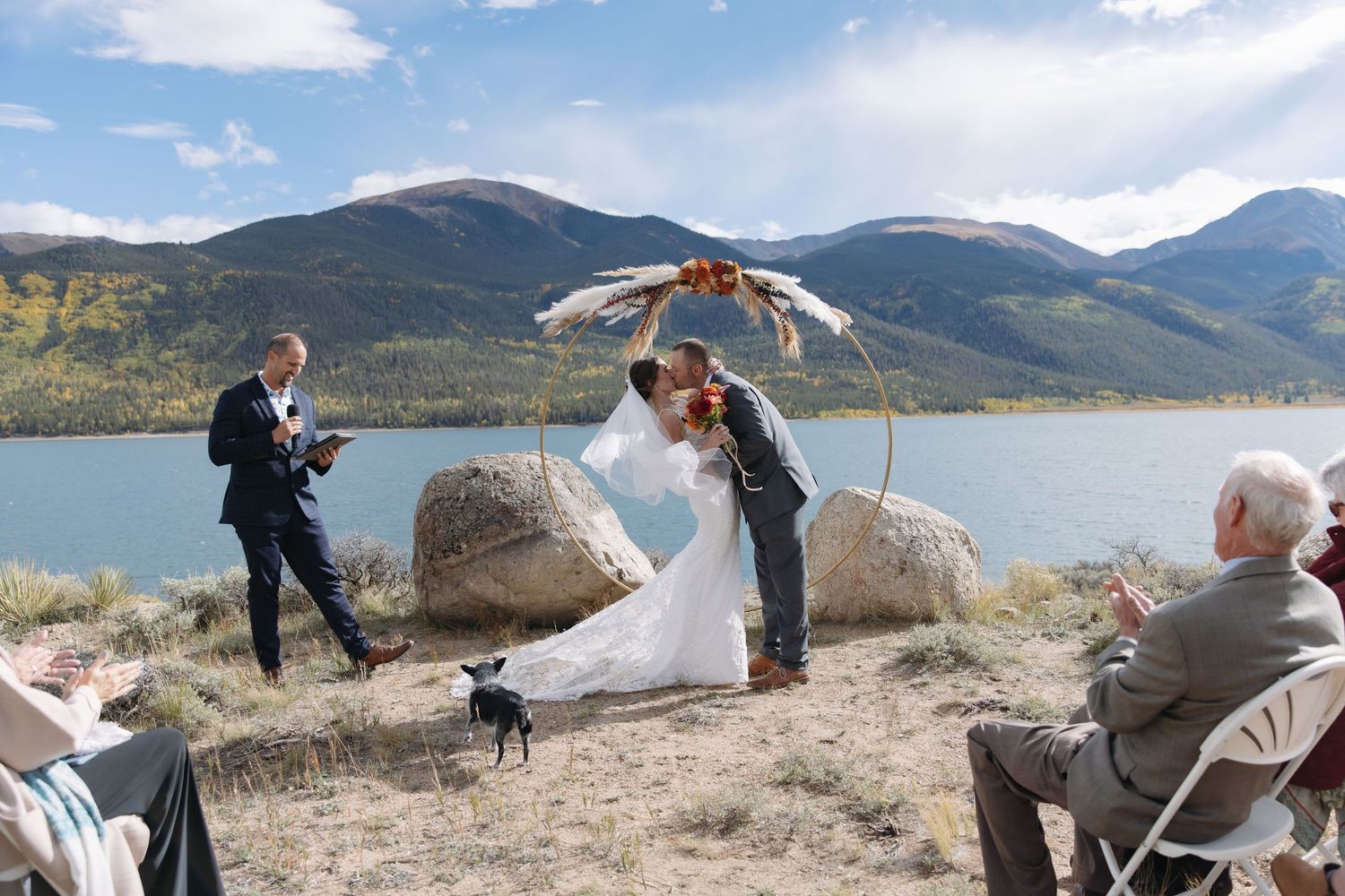 Couple shares first kiss at outdoor lakeside wedding ceremony beneath feathered arch with stunning mountain scenery.