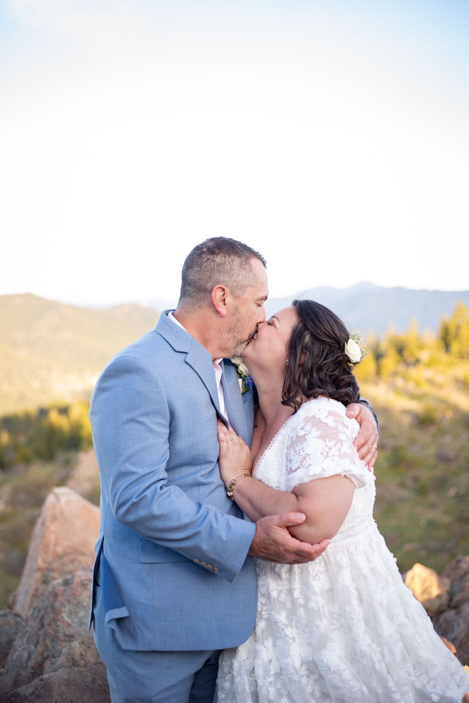 Newlyweds sharing first kiss on rocky mountain overlook, bride in lace gown and groom in blue suit.