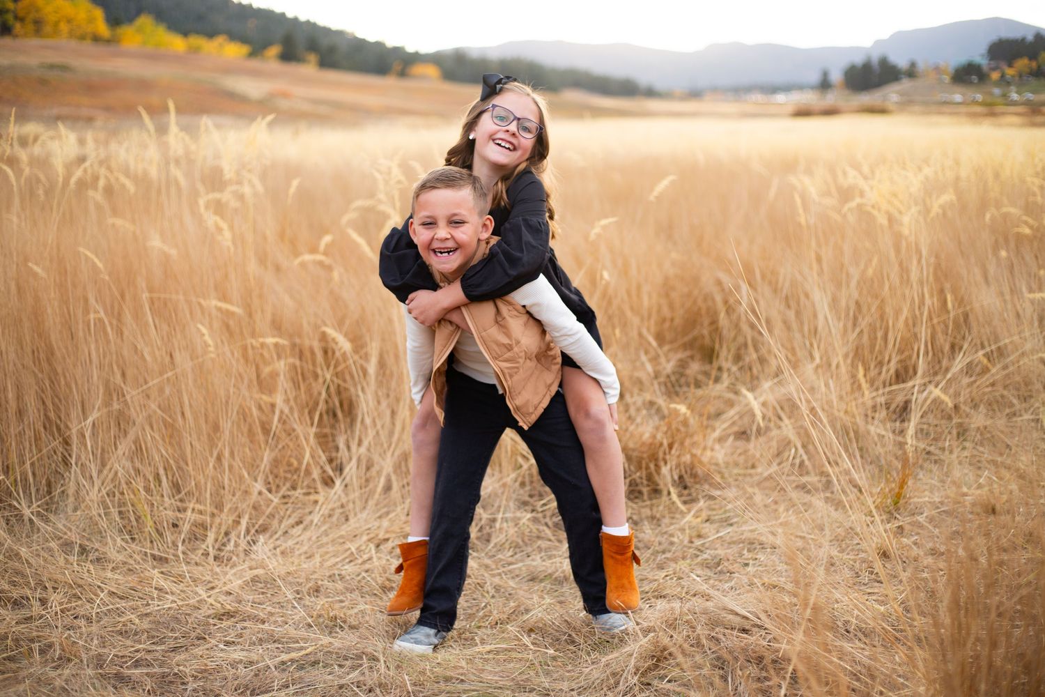 Two children playing piggyback in a golden wheat field with mountain backdrop in autumn.