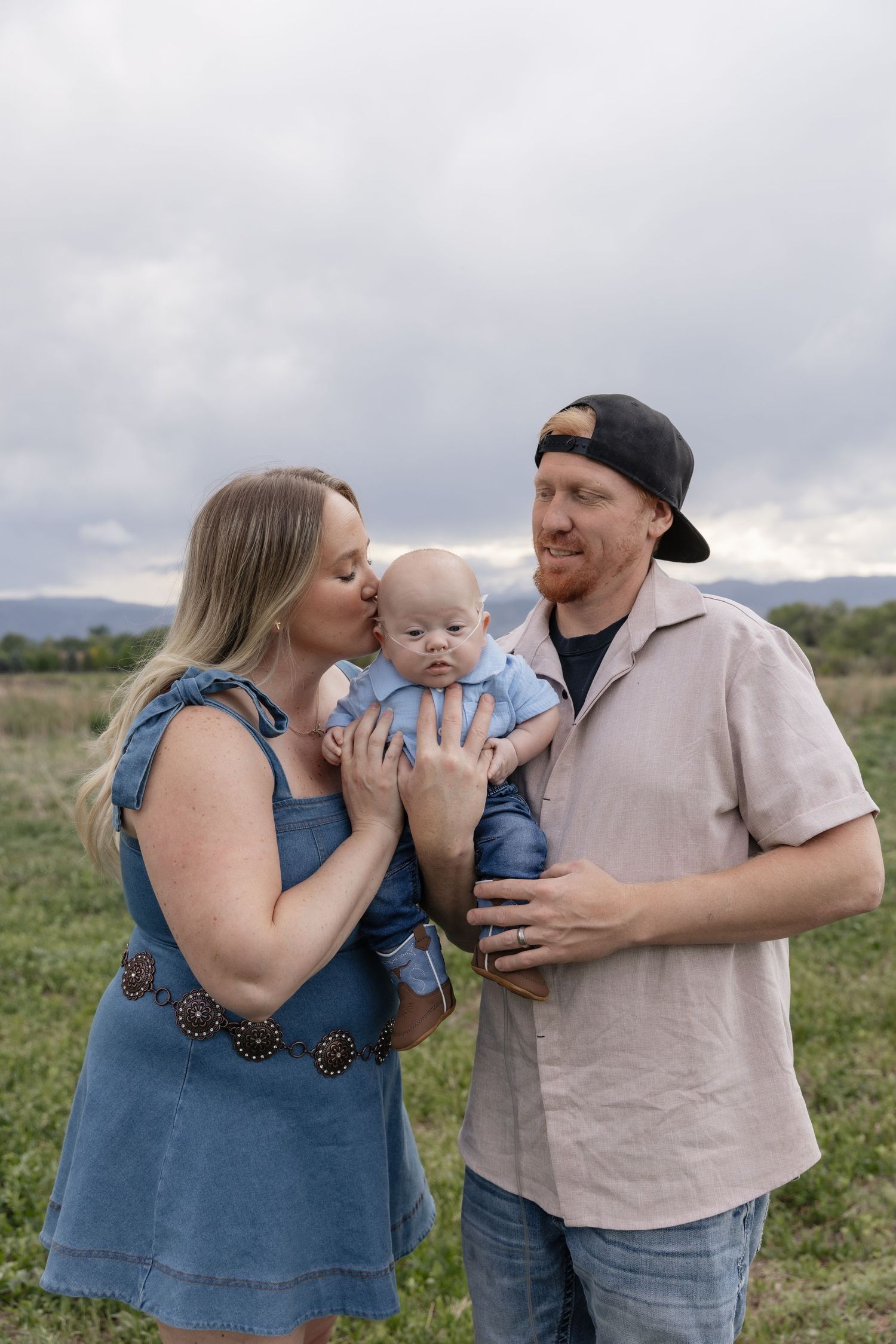 A young family poses outdoors, mother kissing baby in denim, father in cap holding child, green field and cloudy sky behind.