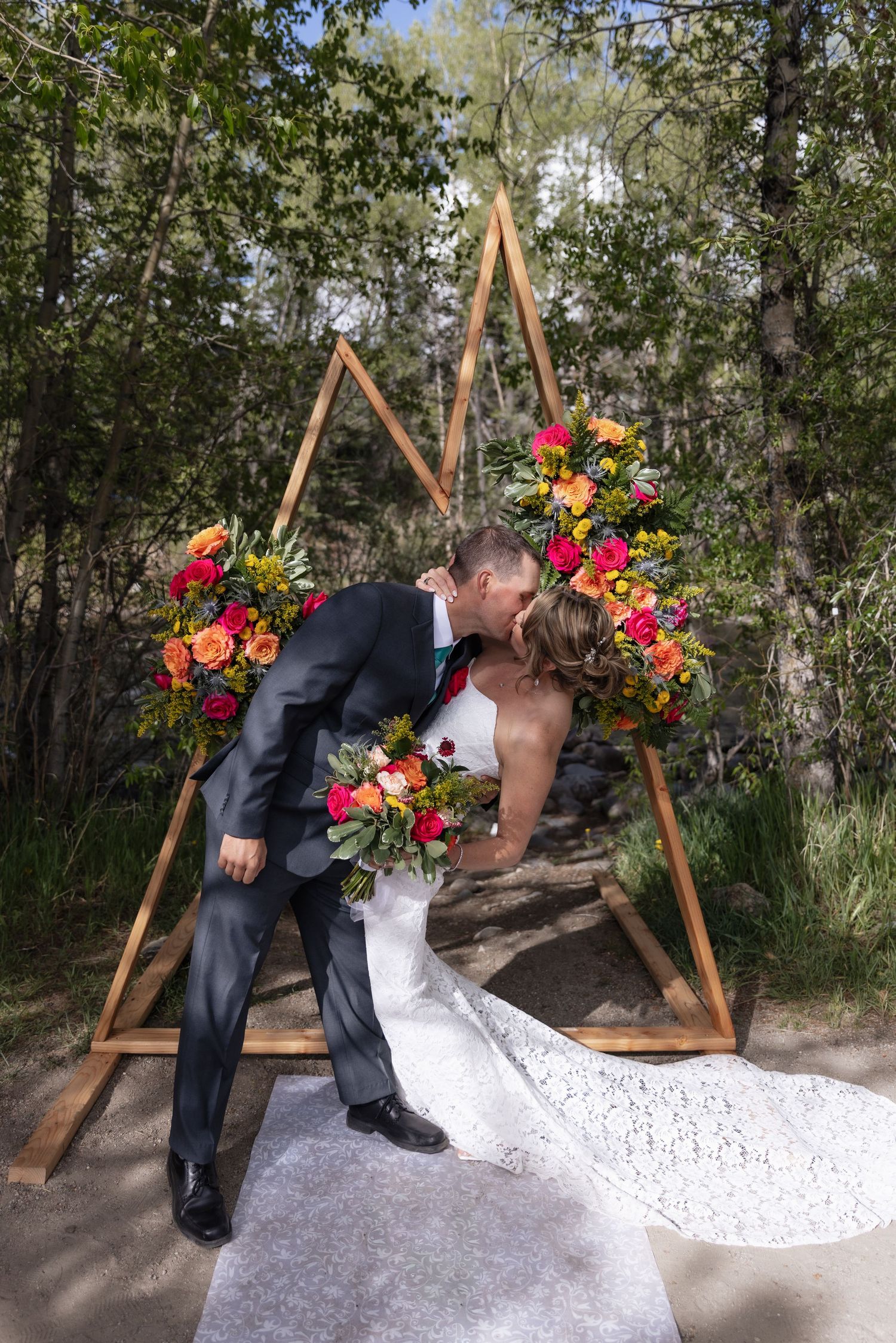 Bride and groom share a romantic kiss under a wooden triangular arch adorned with vibrant red and orange floral arrangements outdoors.