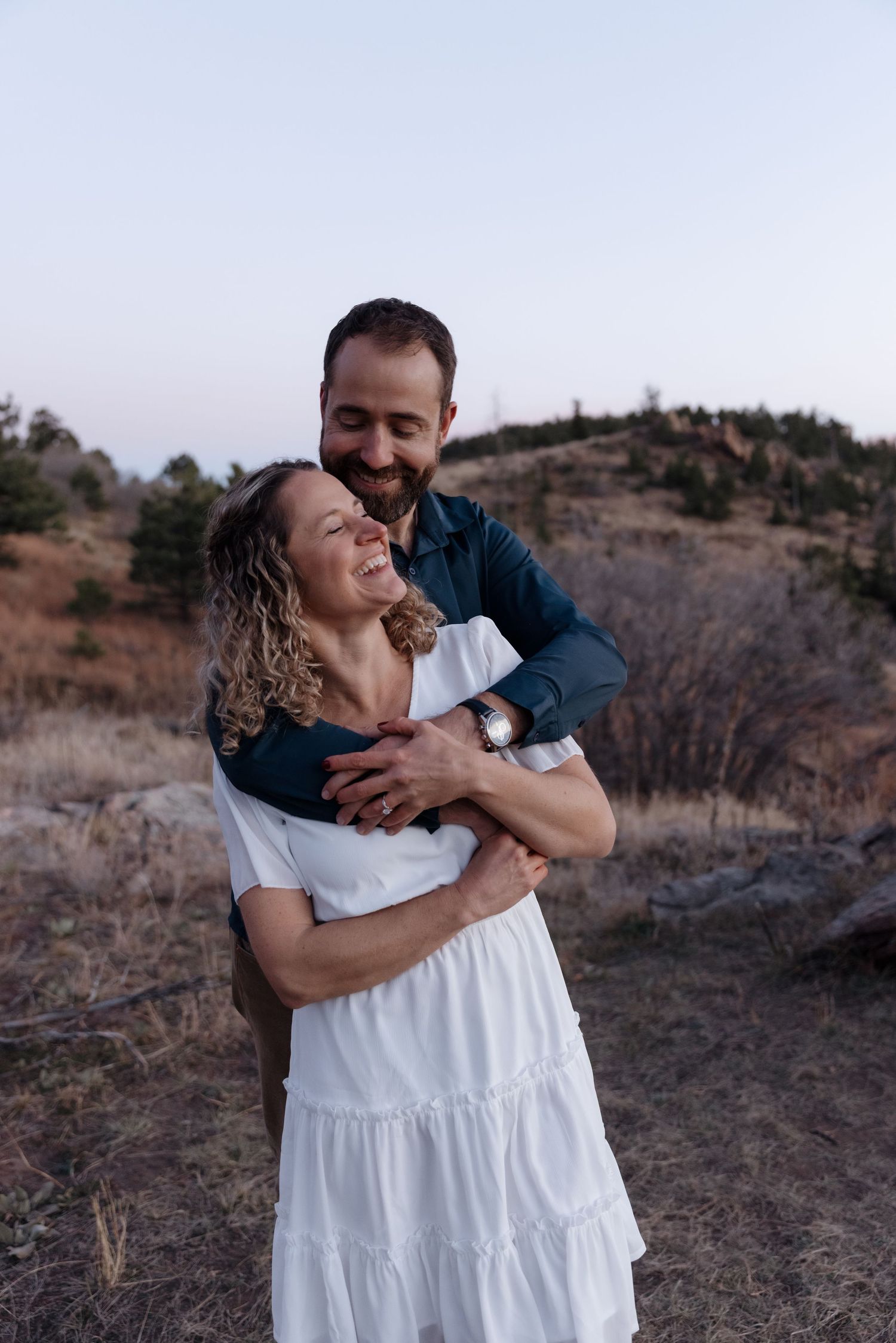 Man hugging woman from behind in a dry meadow, both smiling during outdoor photo session.