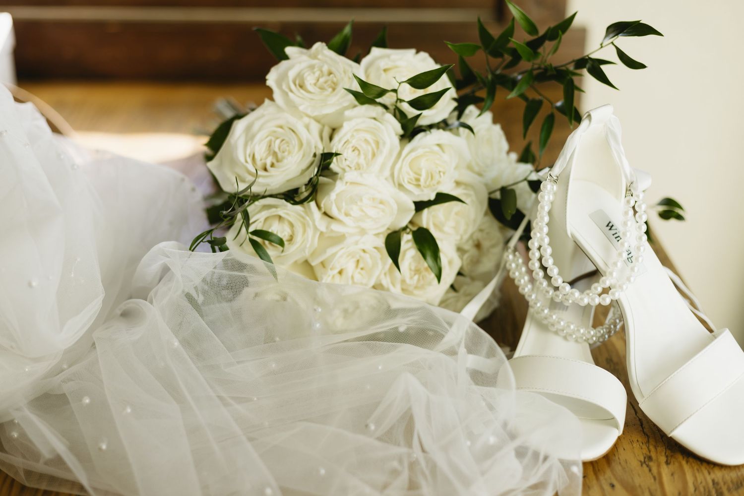 White rose bridal bouquet with pearl-adorned heels and veil on a wooden surface.