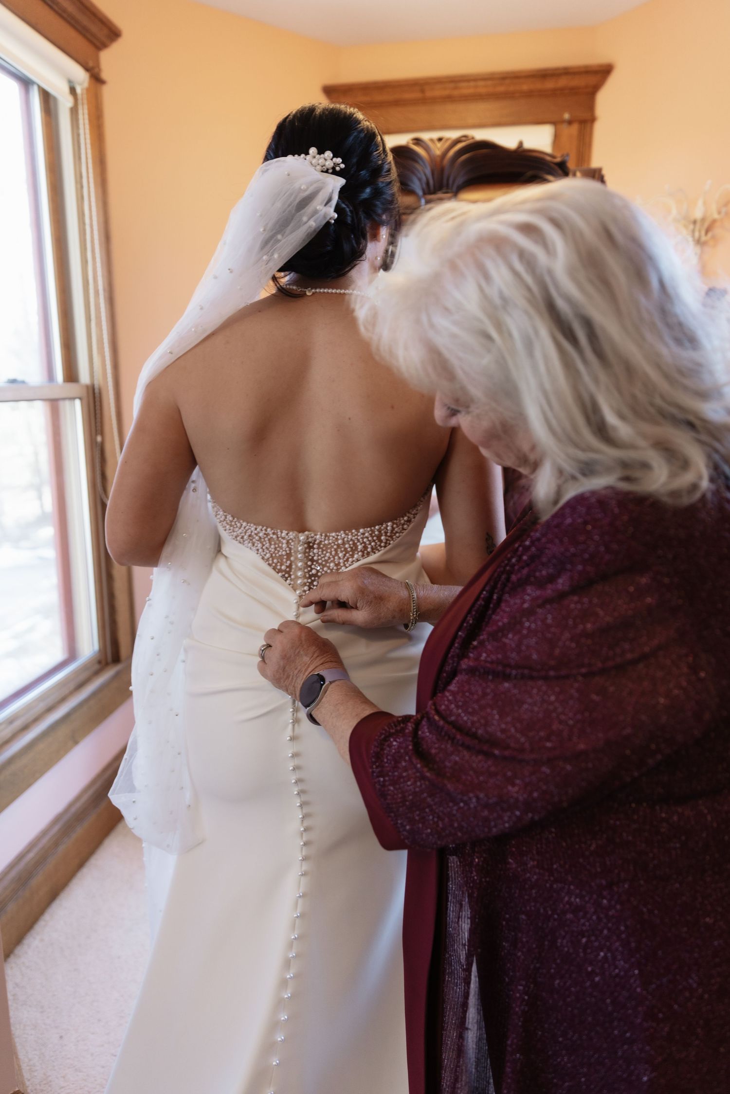 Mother helping bride button the back of her beaded wedding gown near a bright window.