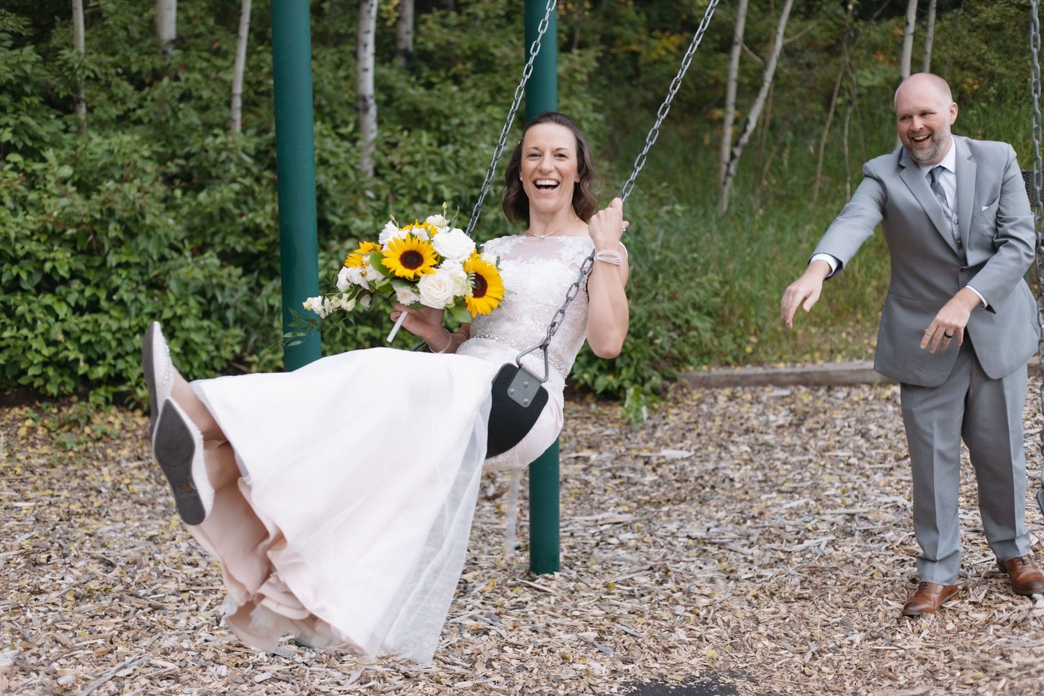 Laughing bride in white dress swings while groom pushes her at outdoor playground.