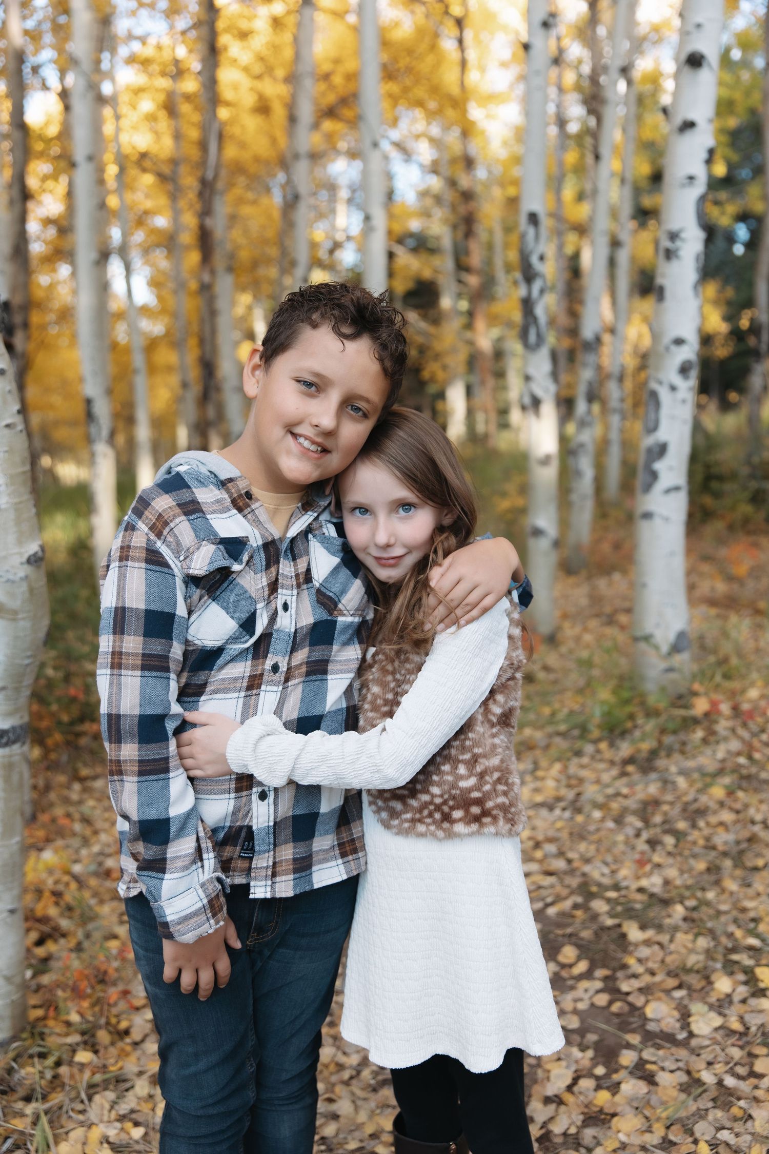 Two children hug and smile among golden aspen trees in fall.